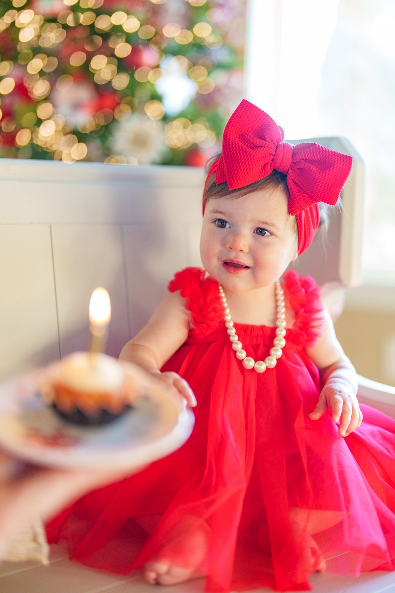 Baby girl in red dress and bow headband receiving a birthday cupcake with lit candle near Christmas tree.