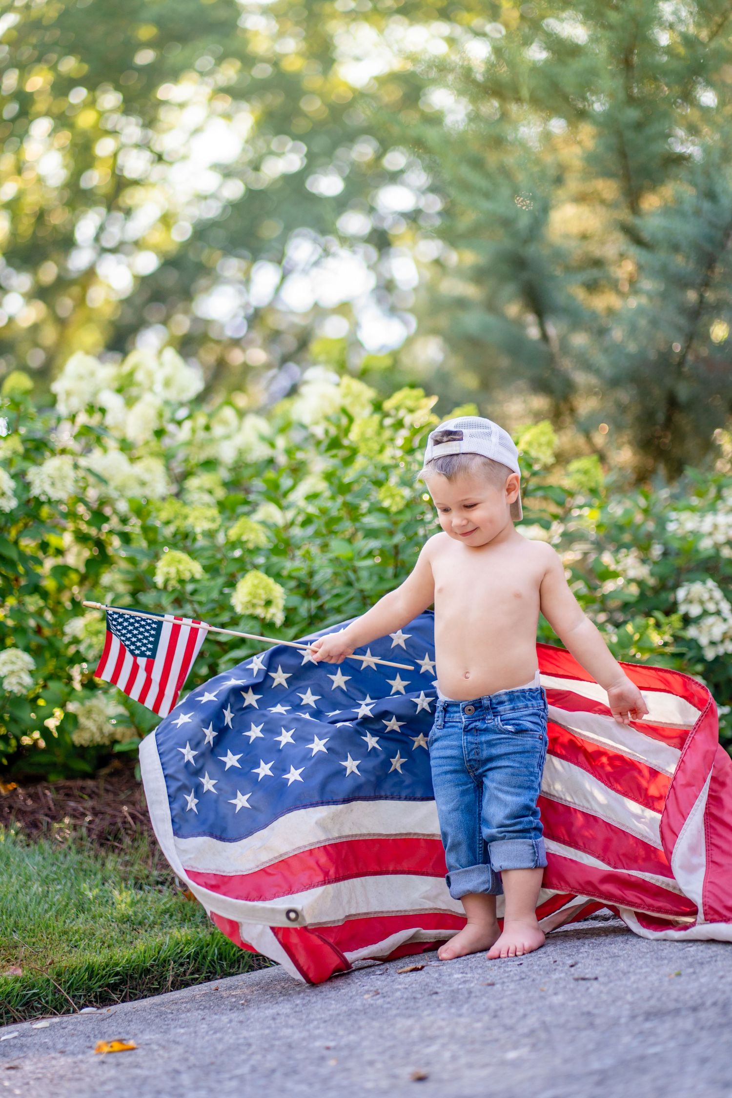 Shirtless toddler boy wrapped in American flag holding small USA flag outdoors.