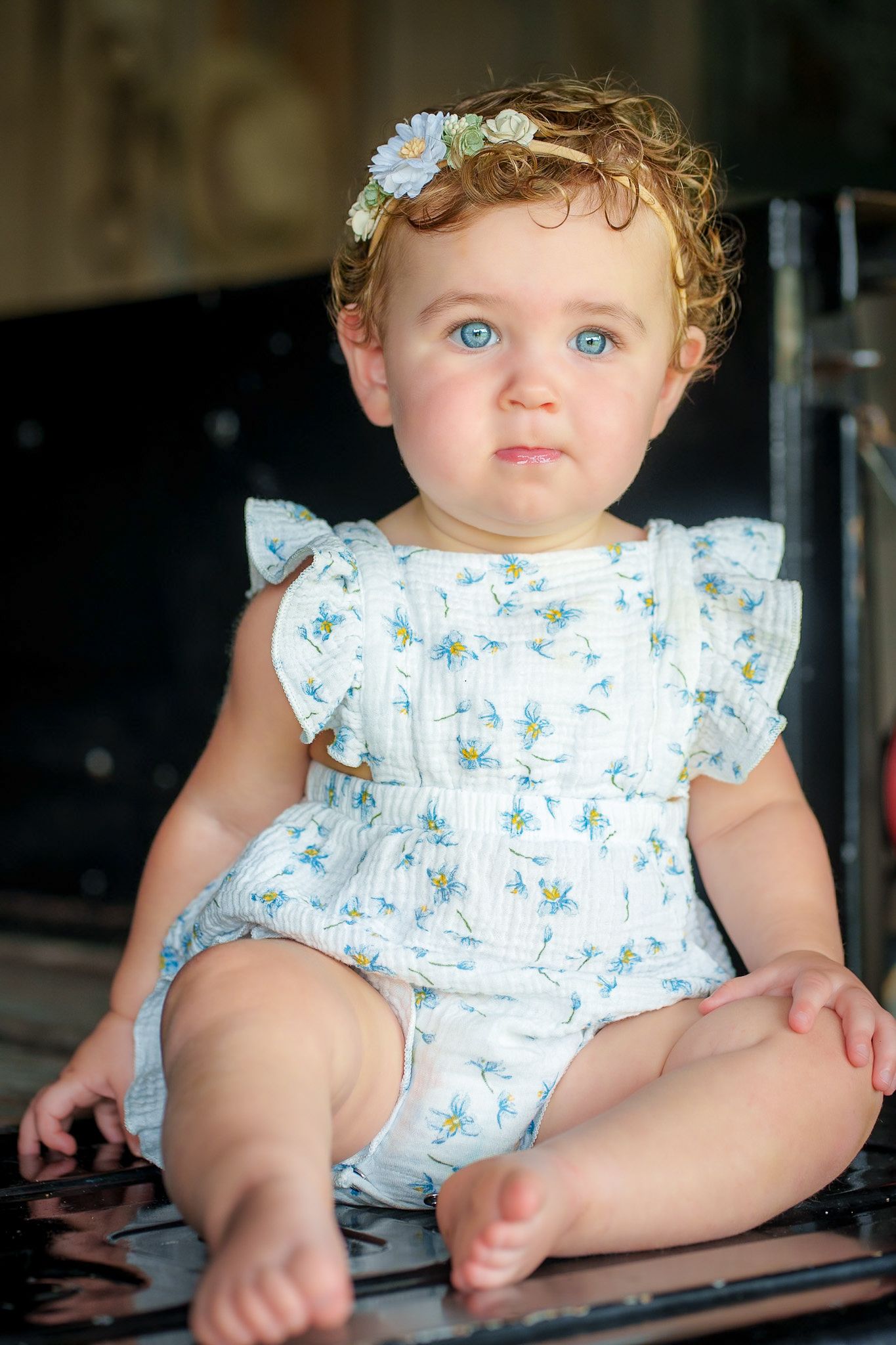 A blue-eyed baby girl in a floral romper and daisy headband sits and gazes forward in soft light.