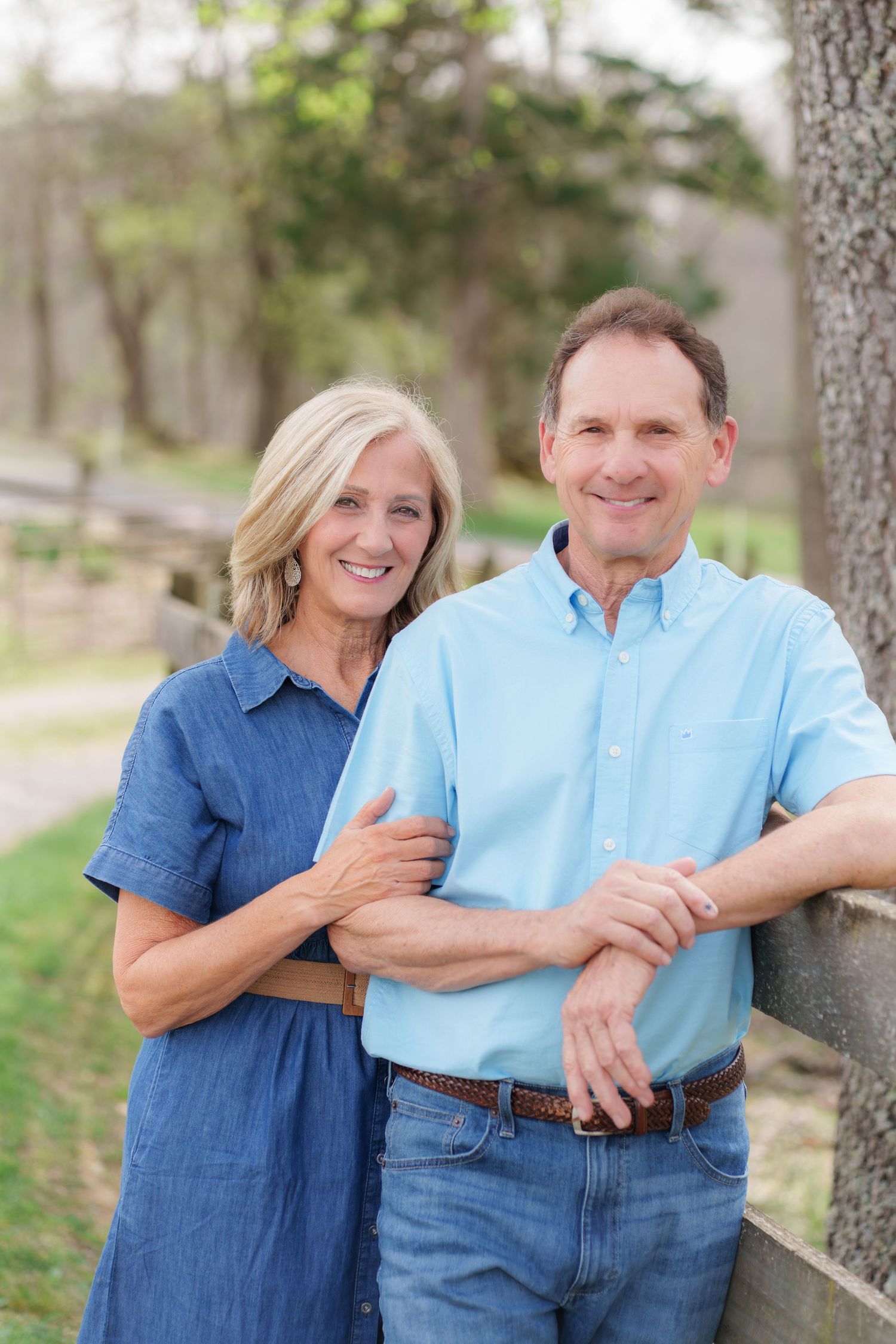 Smiling middle-aged couple posing outdoors near a wooden fence and tree in a lush green park setting.