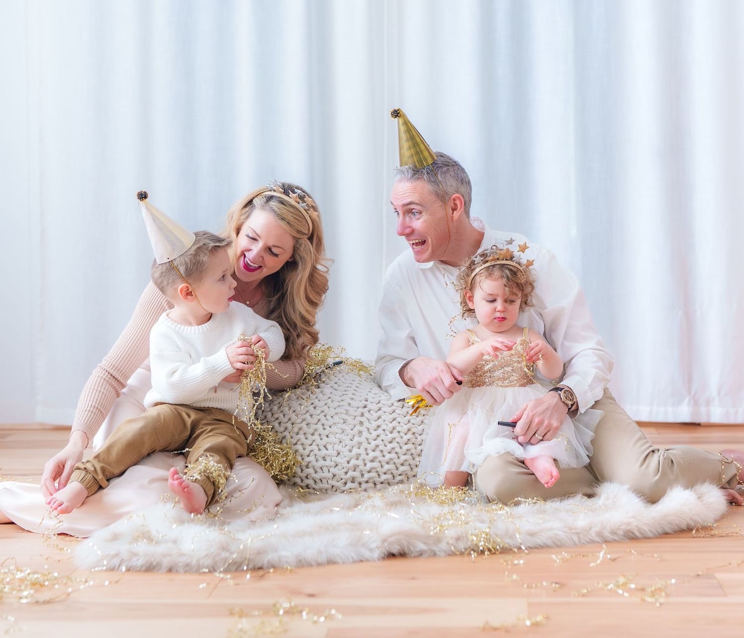 Joyful family of four wearing gold party hats celebrating New Year together on a fur rug.