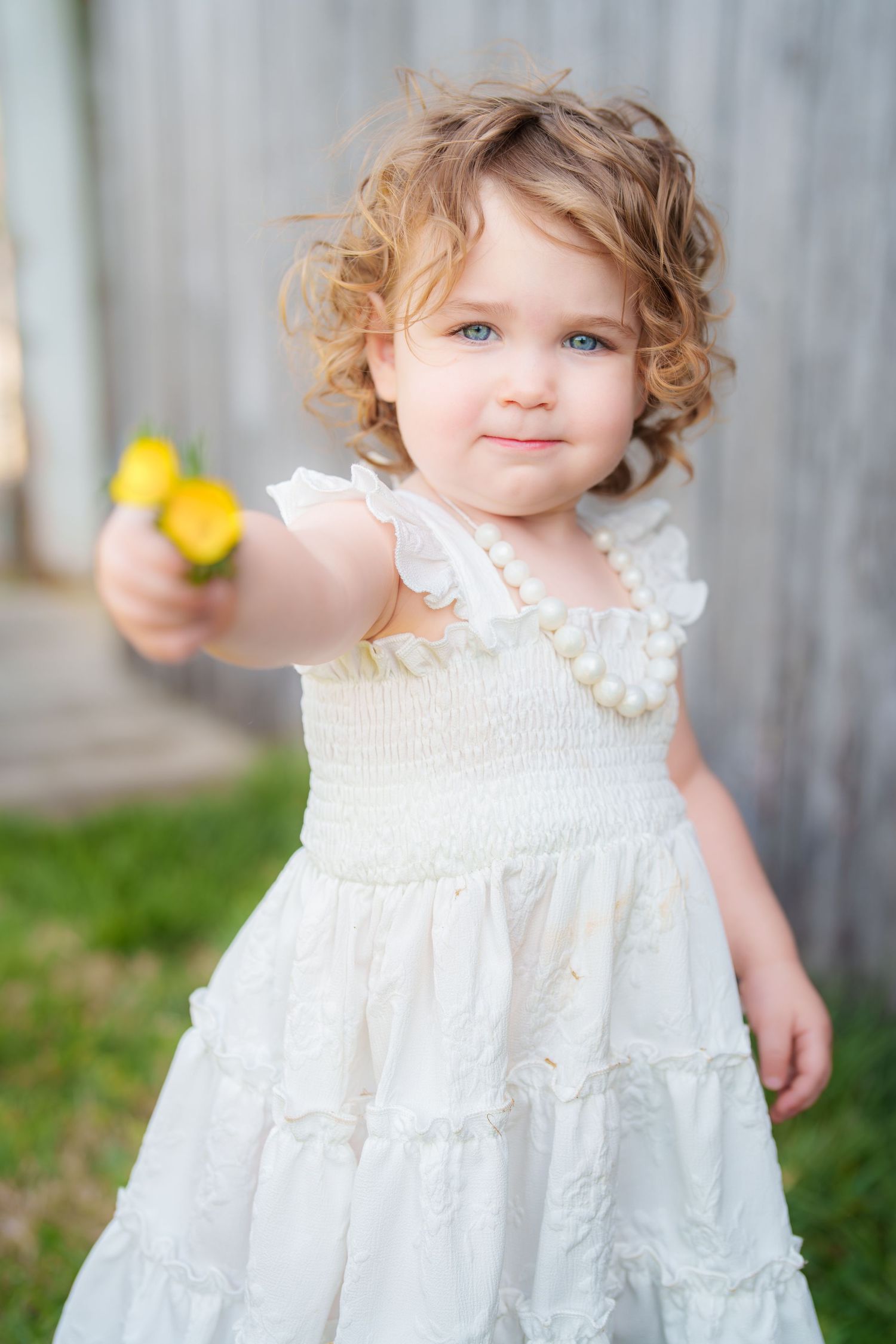 Adorable toddler girl with curly hair in white dress holding yellow flower outdoors.