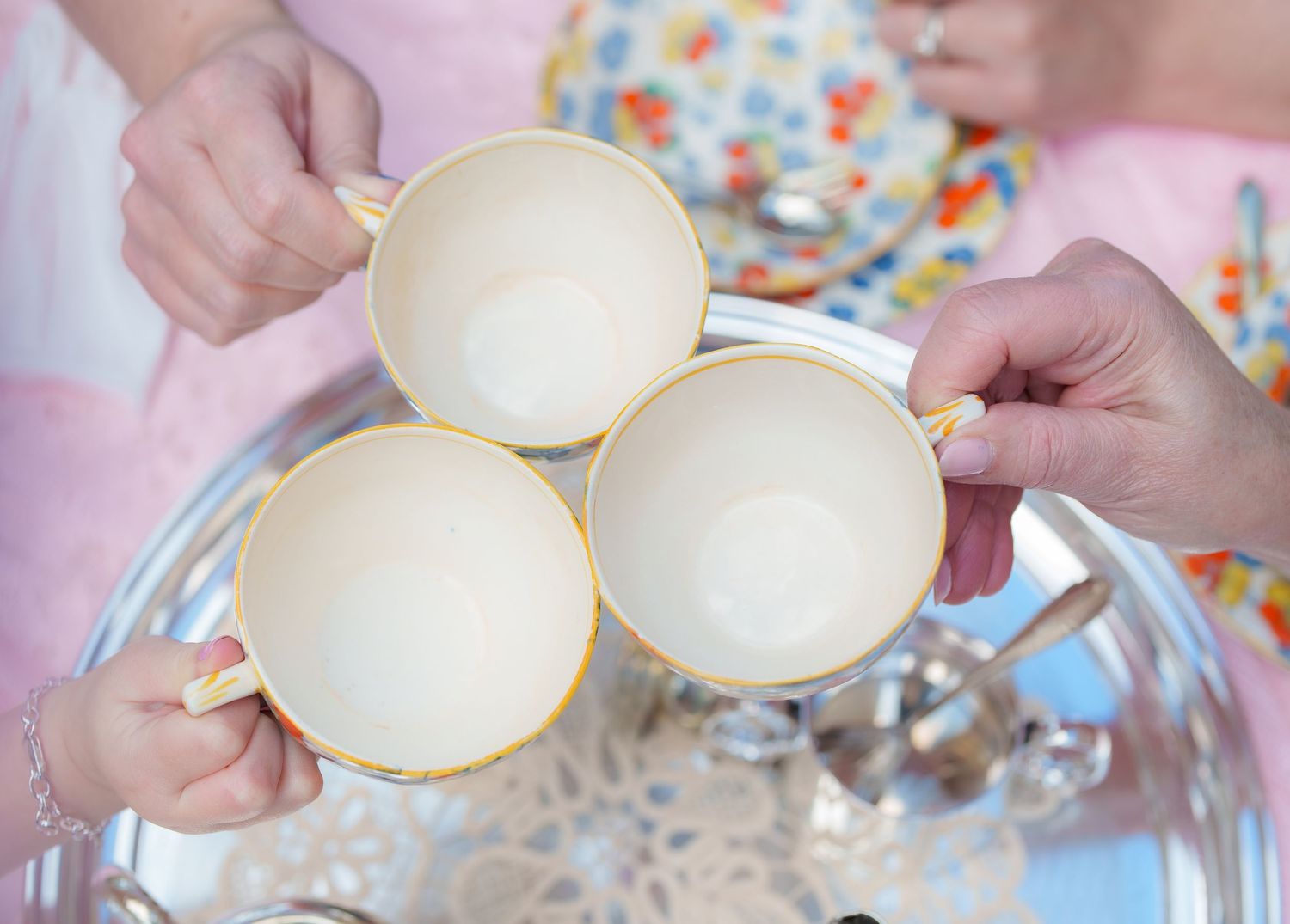 Three hands clink yellow-rimmed teacups together over a silver tray at a tea party.
