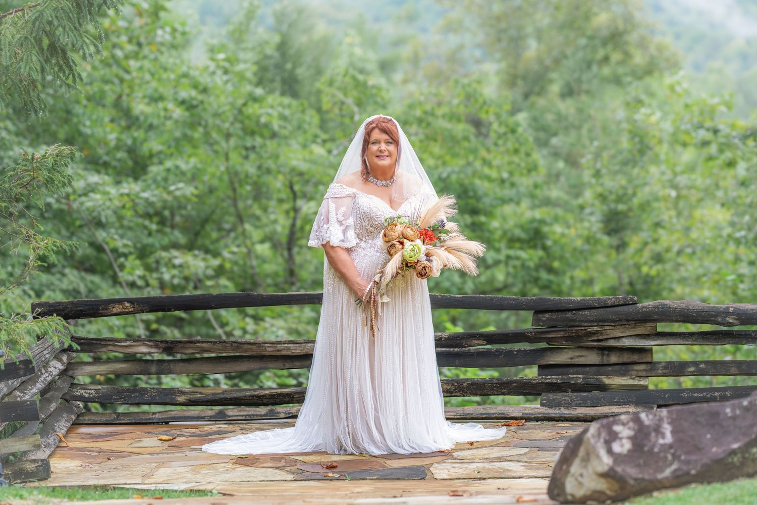 Bride in white lace gown holding bouquet on wooden bridge surrounded by lush green forest.