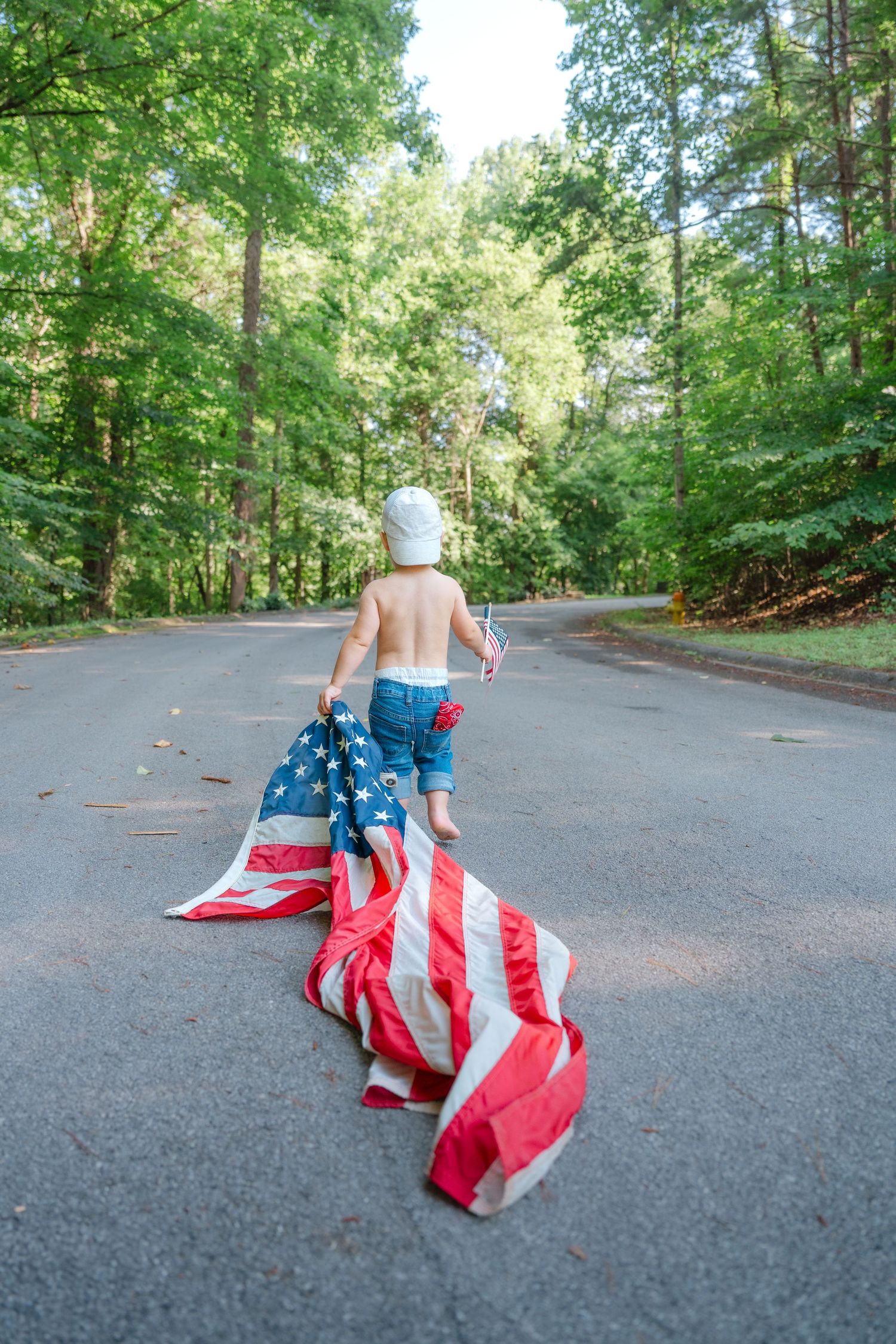 Young child running down a tree-lined road dragging a large American flag behind them.