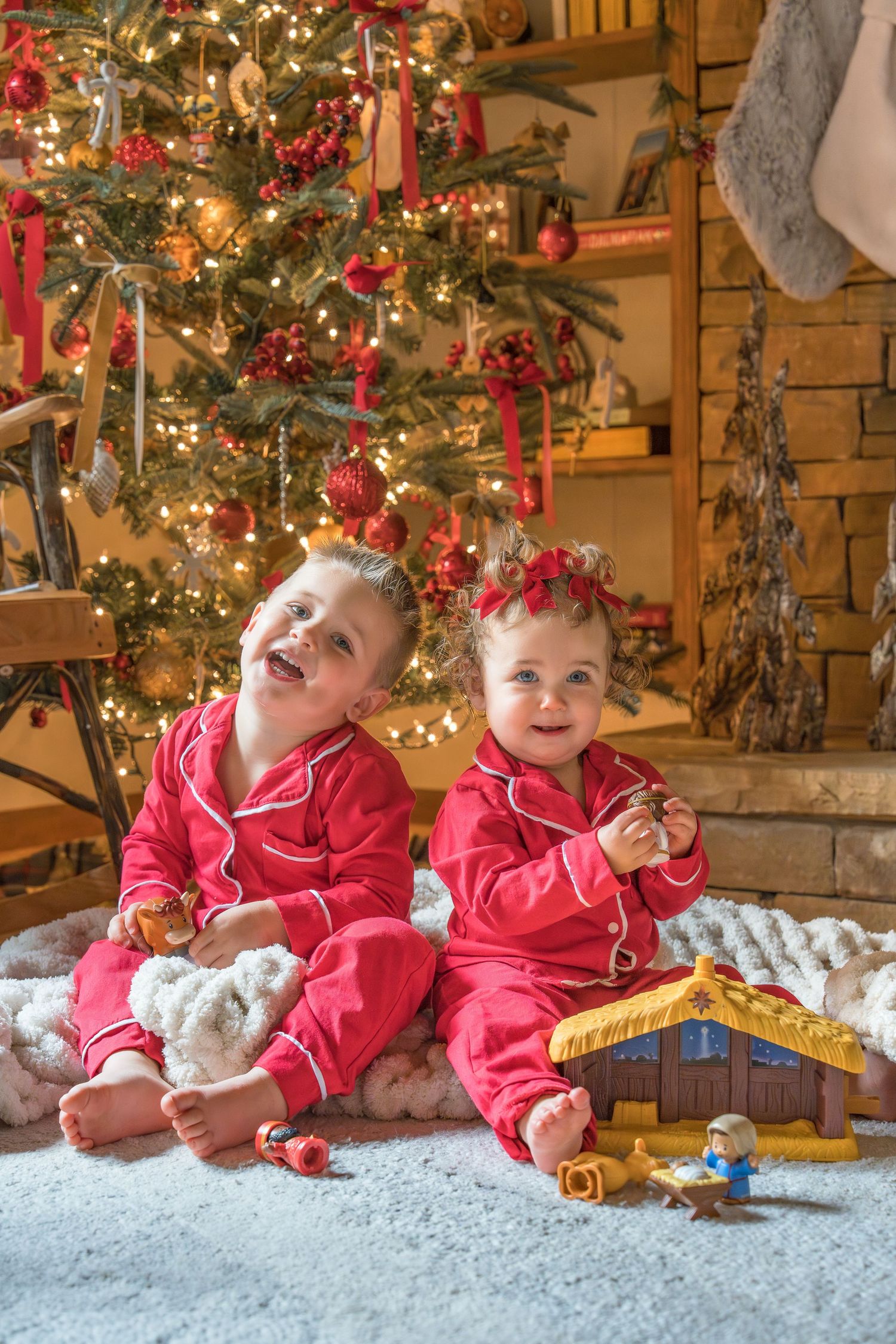 Two babies in red Christmas pajamas sit smiling before a decorated tree with a nativity toy set.
