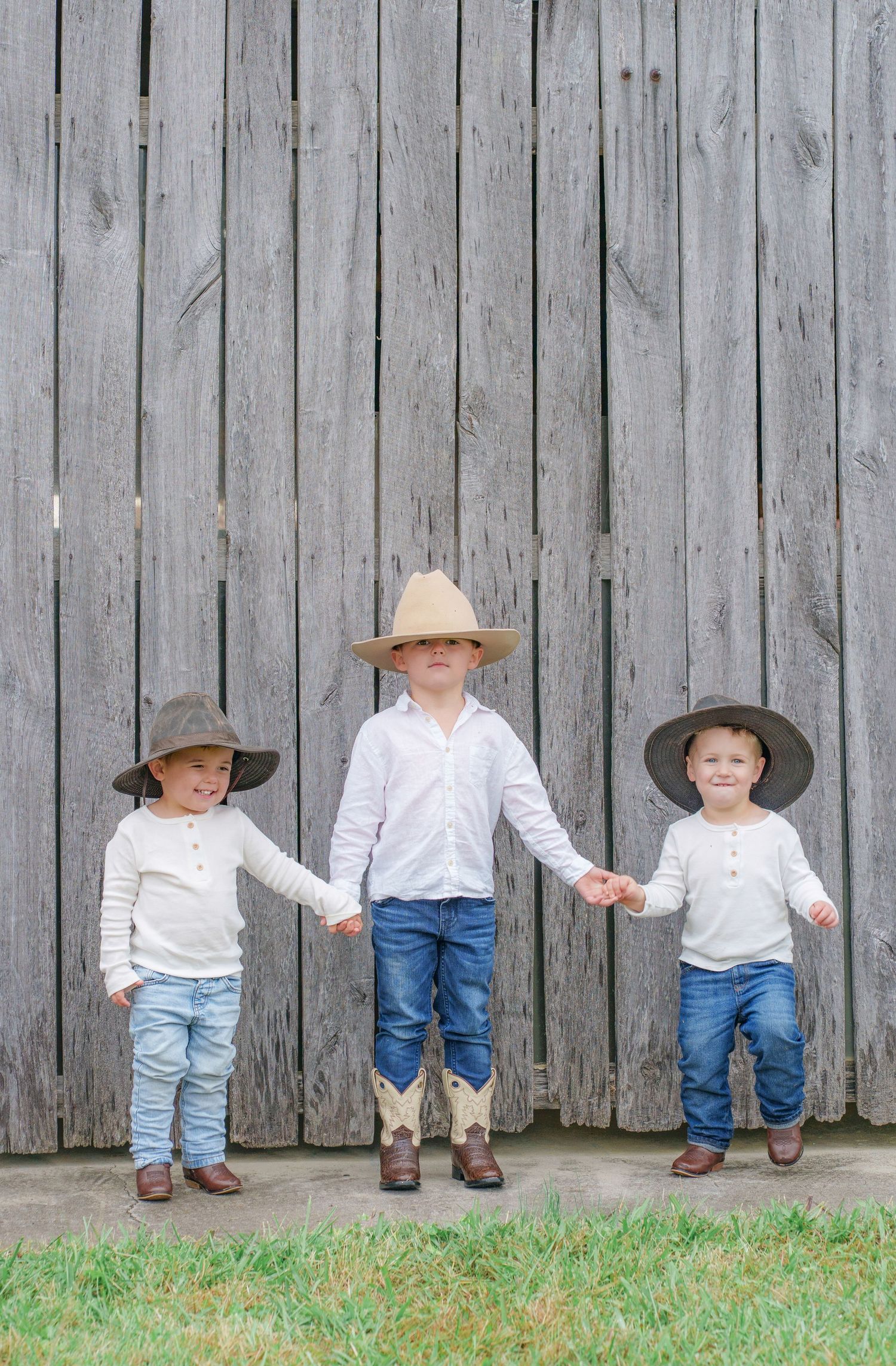 Three young boys in white shirts, jeans, and cowboy hats hold hands in front of a weathered gray wooden barn wall.