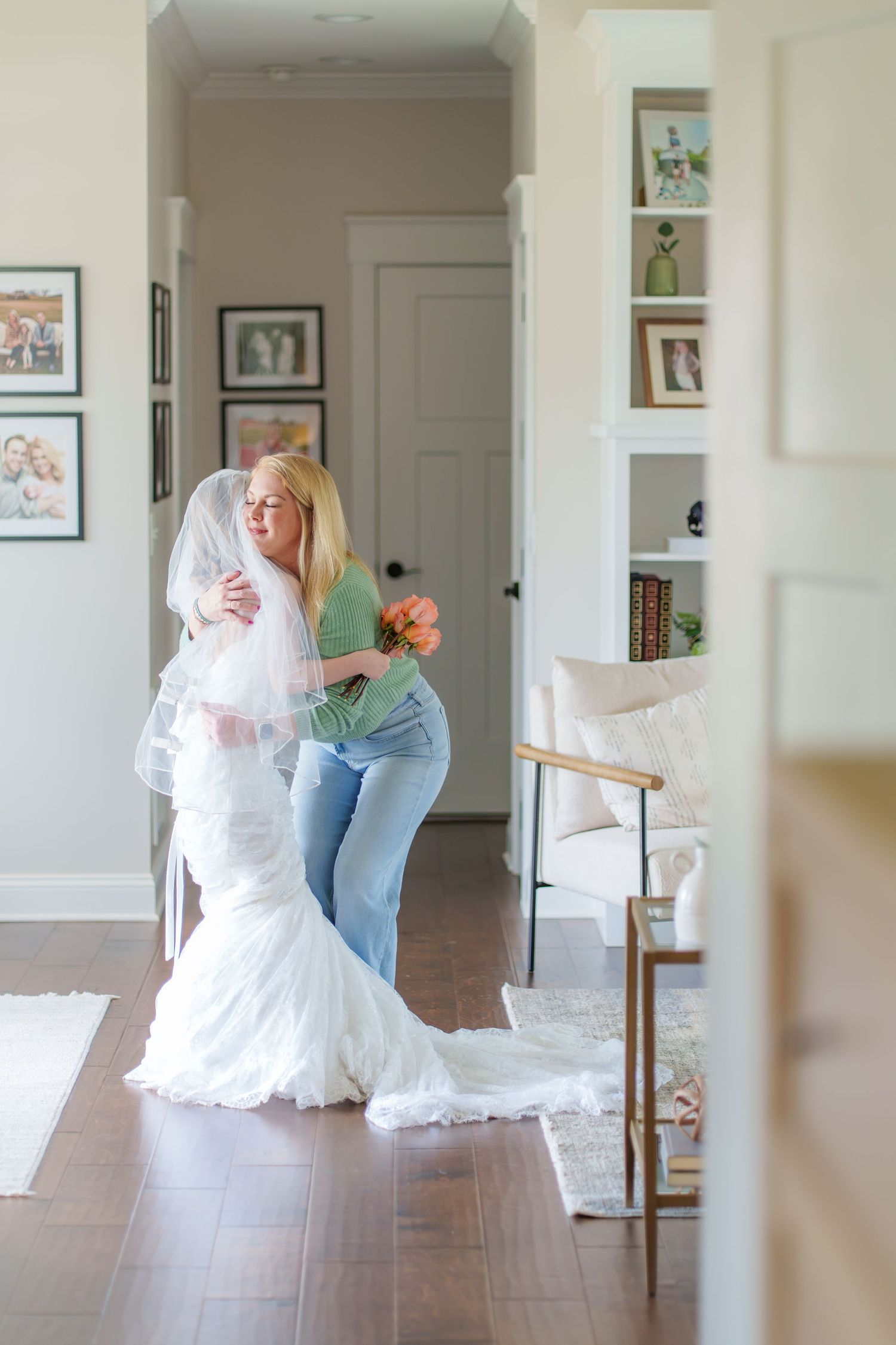 A blonde woman in green embraces a bride in a wedding gown inside a bright home.