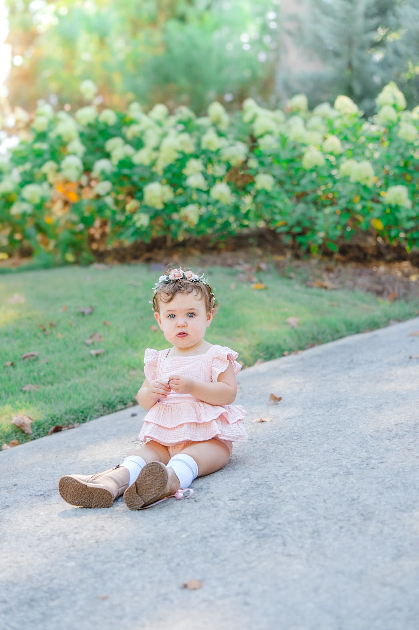 Baby girl in pink ruffled outfit and floral crown sitting on a pathway surrounded by green bushes.