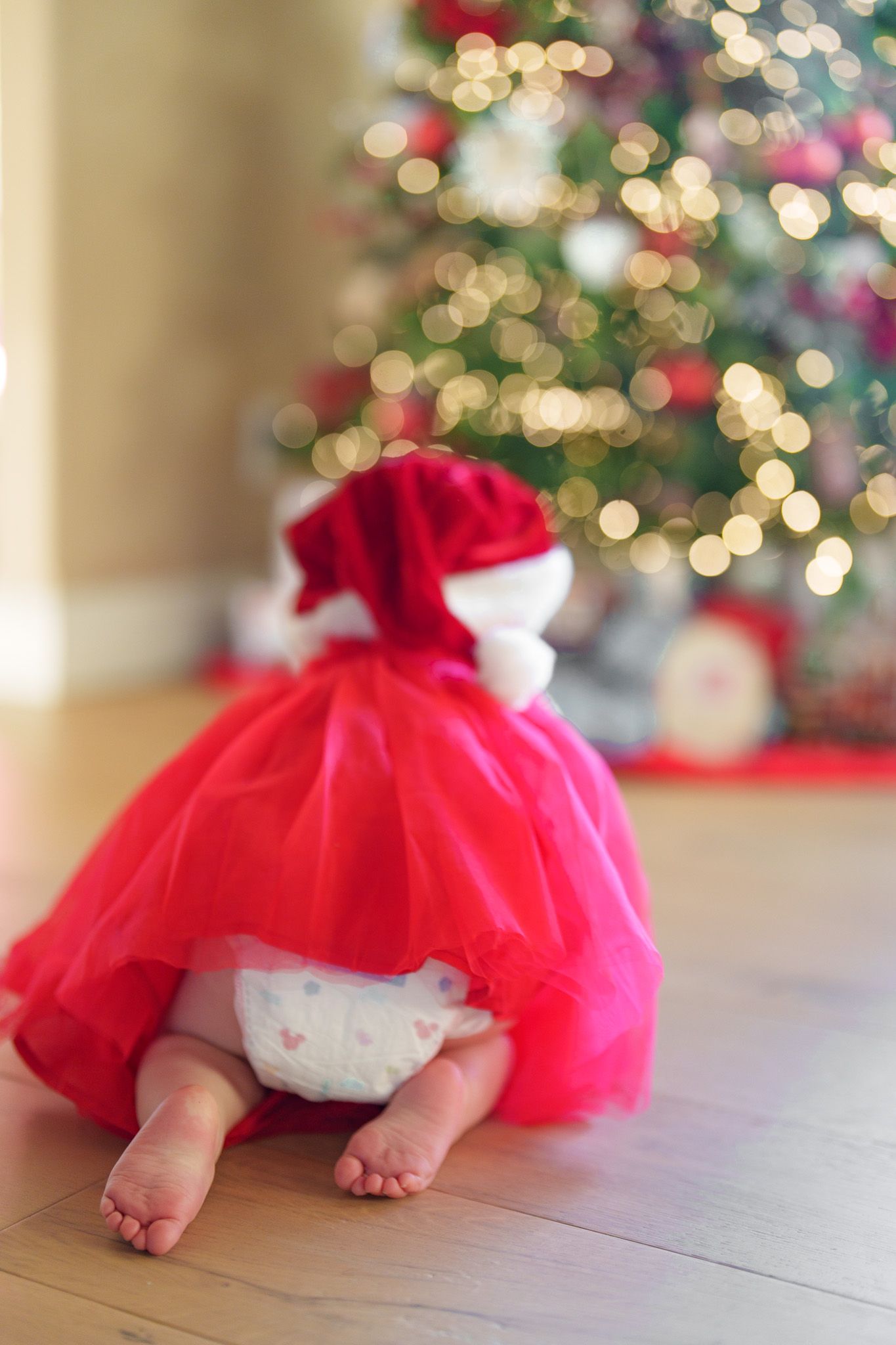 Baby in red tutu dress and Santa hat sitting on floor in front of decorated Christmas tree.