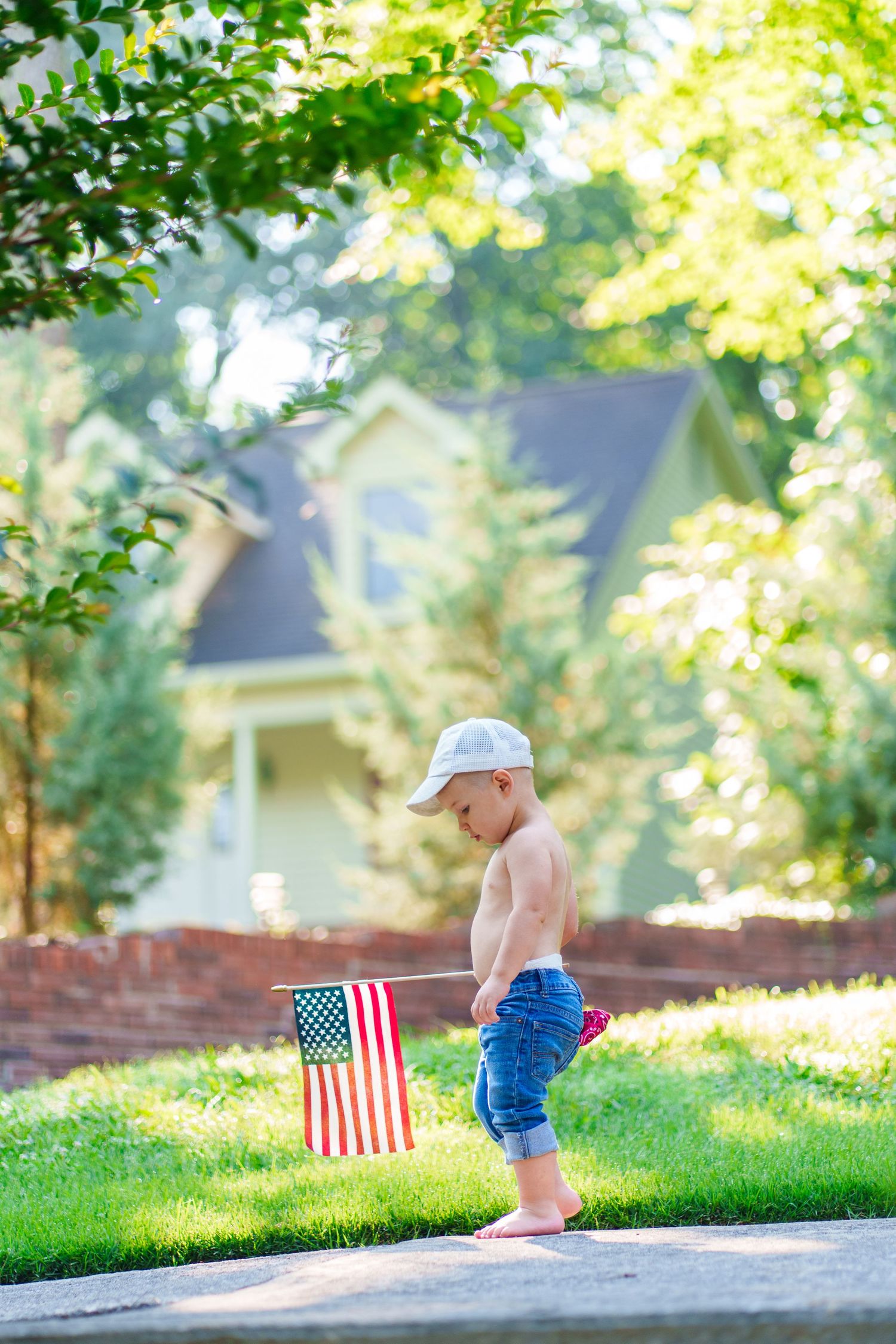 A shirtless toddler in jeans and cap holds an American flag while walking on a sunny sidewalk.
