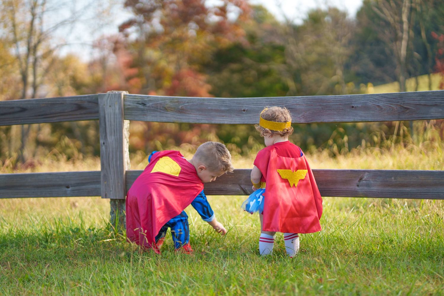 Two children in superhero capes crouching by wooden fence in fall outdoor setting.