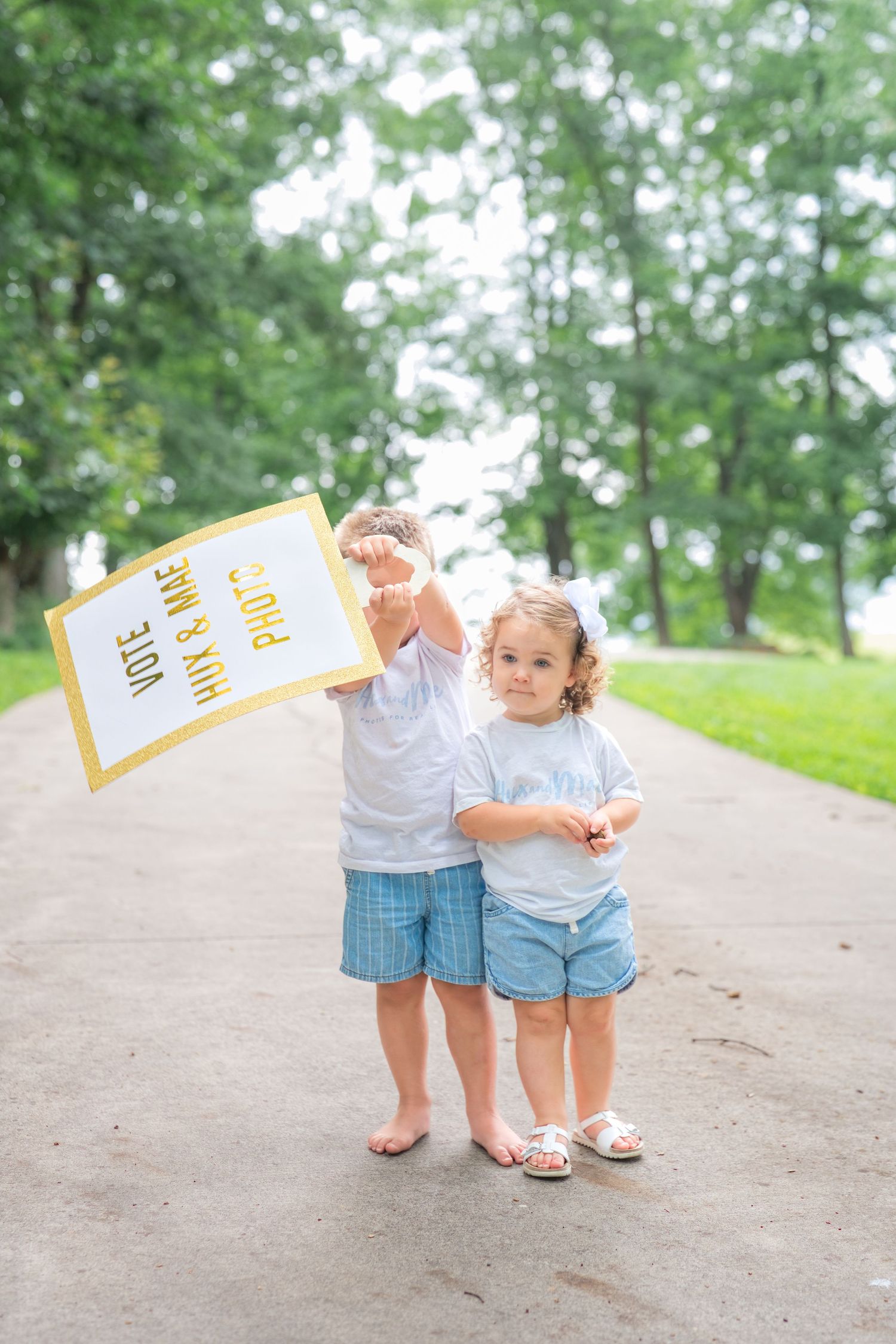 Two young children in white shirts holding a vote sign on a tree-lined path outdoors.