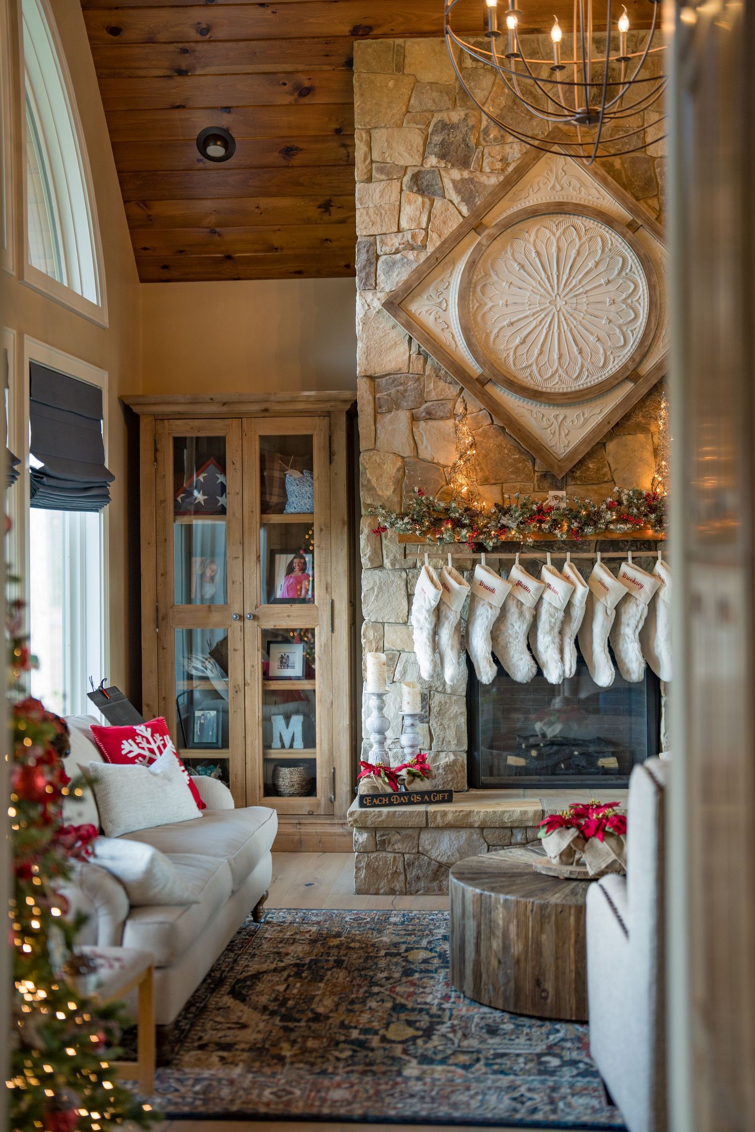 Cozy Christmas living room with stone fireplace, hanging stockings, garland, and decorated tree in rustic cabin setting.