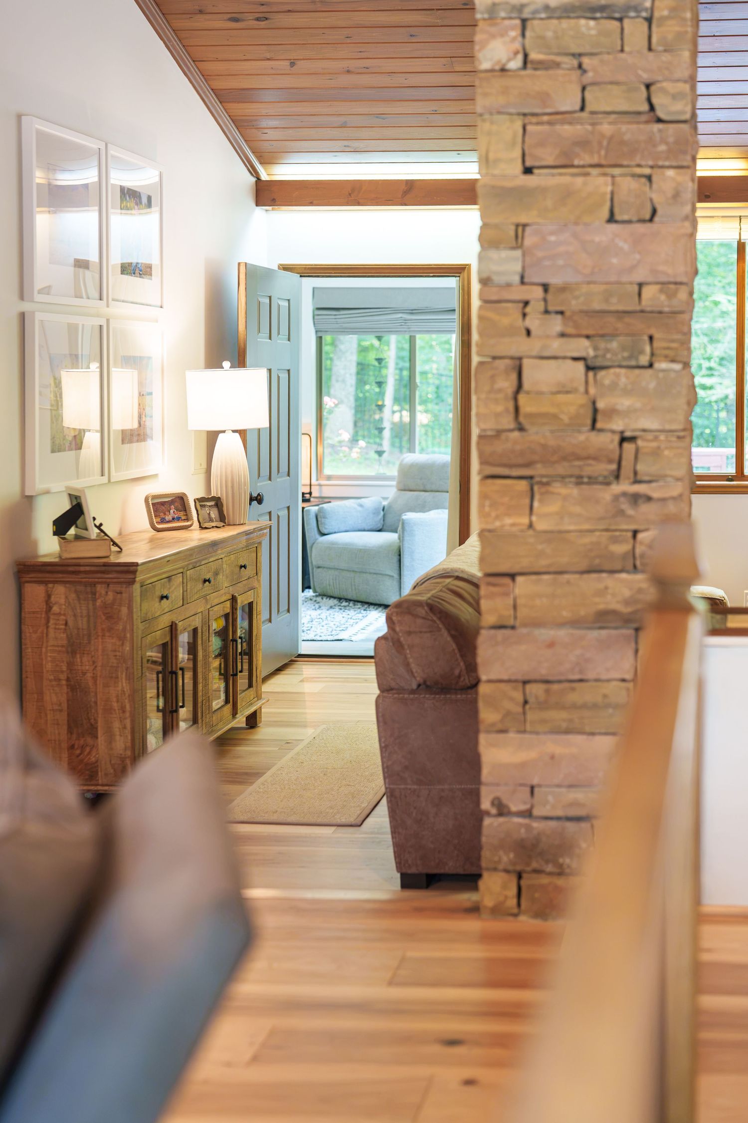 Rustic home interior with stone pillar, wood ceiling, hardwood floors, console table with lamp, and bright doorway beyond.