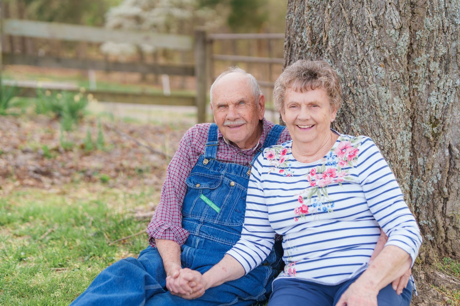 Happy senior couple sitting together against a large tree trunk in a grassy yard.