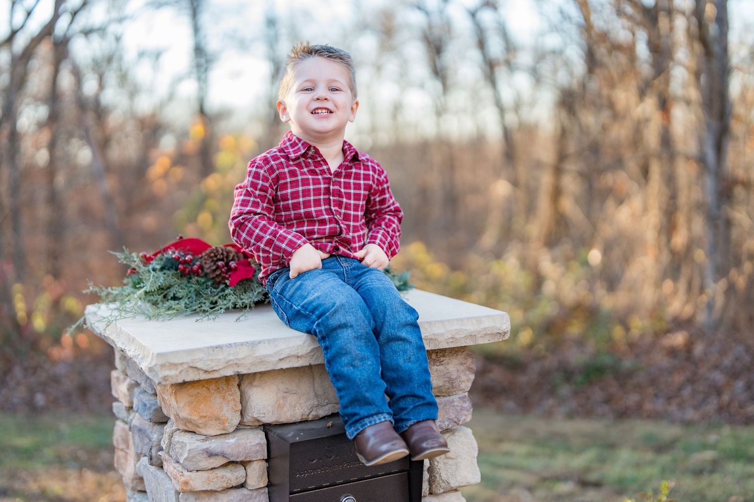 Happy young boy in red plaid shirt sits on stone pillar with holiday poinsettias outdoors.