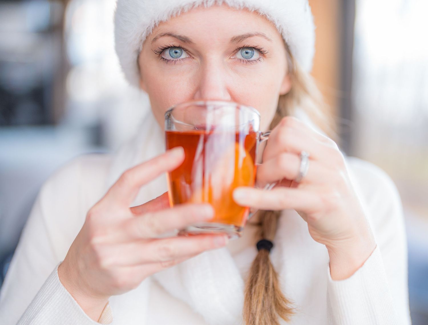 Blonde woman in white knit hat sipping hot tea from a glass cup outdoors in winter.