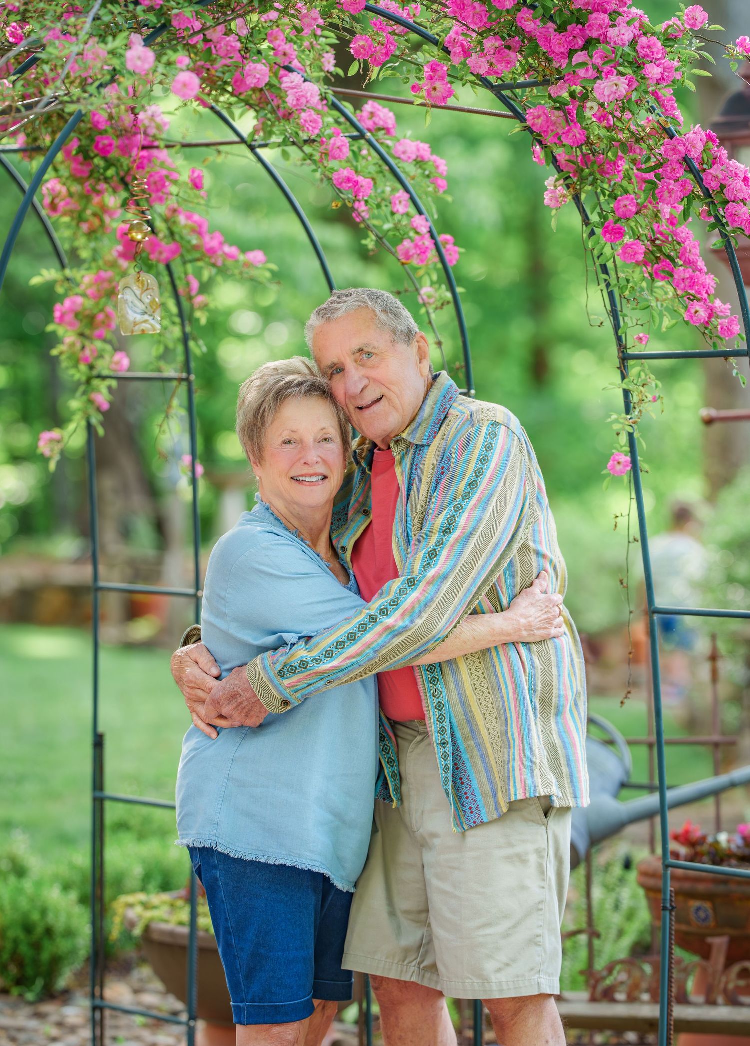 Happy senior couple hugging under a beautiful pink rose arch in a lush green garden.