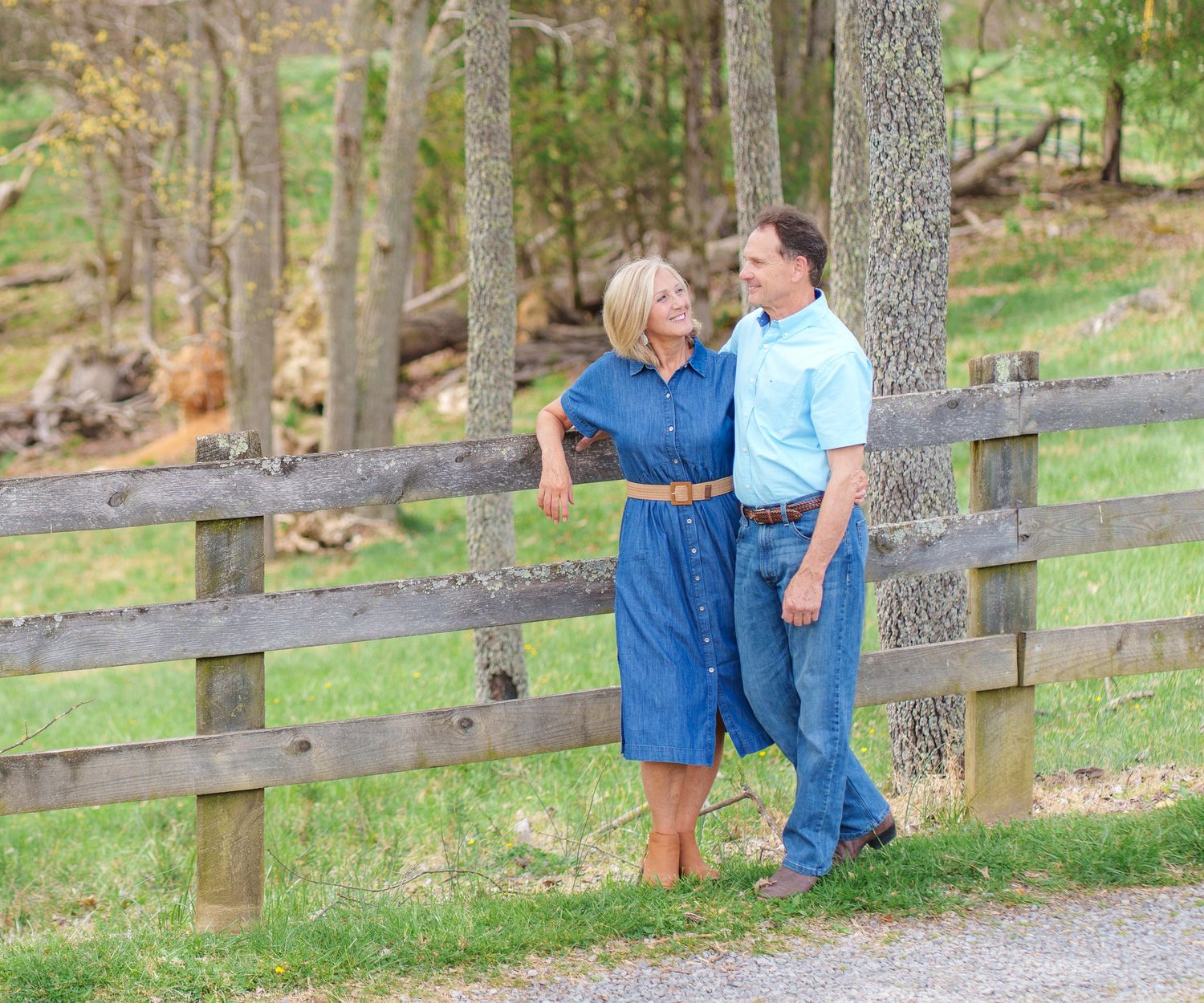 Happy couple leaning against a rustic wooden fence surrounded by trees in a scenic outdoor setting.