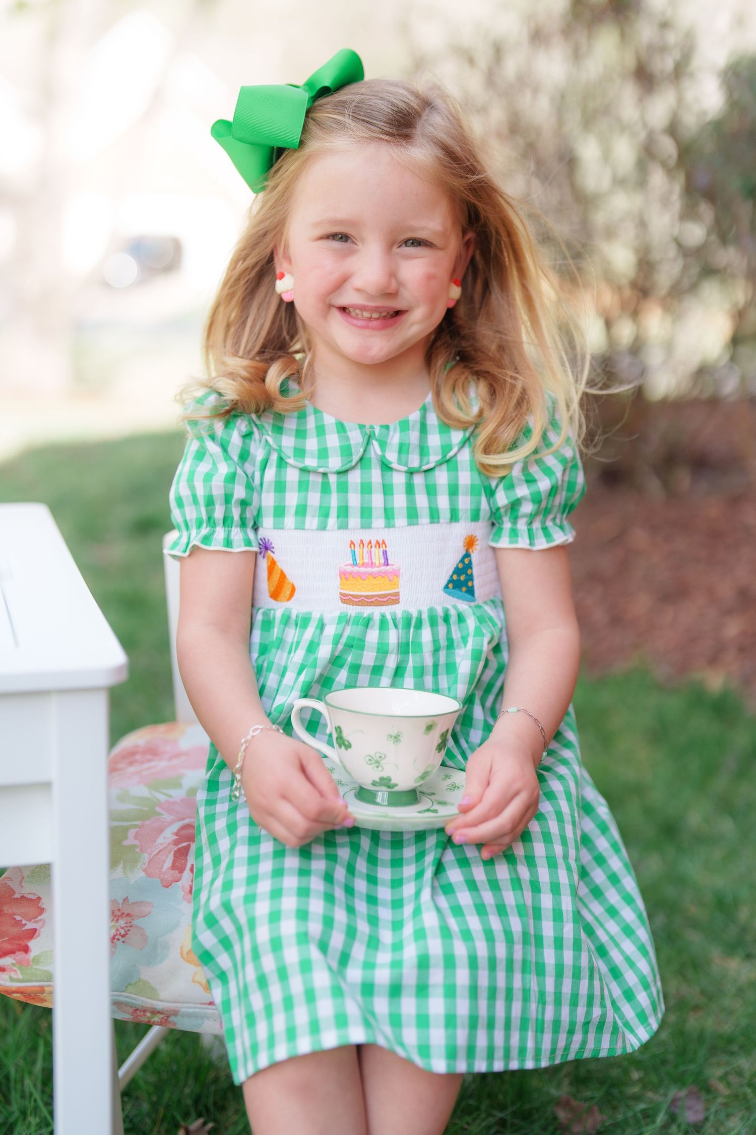 A cheerful girl in a green gingham birthday smocked dress holds a teacup at an outdoor birthday tea party.