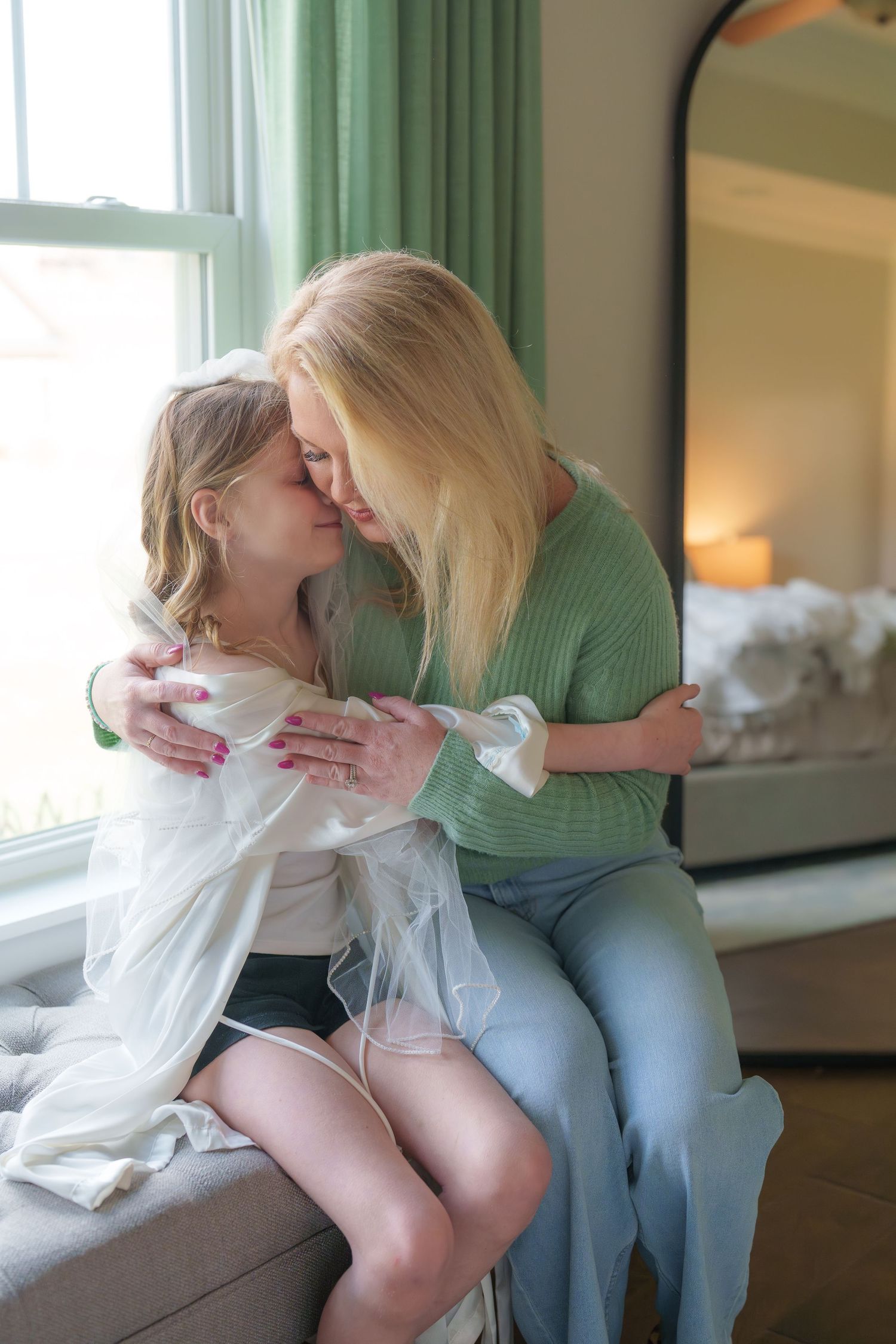A blonde woman and a young girl in a wedding veil share a tender nose-to-nose embrace by a sunlit window.