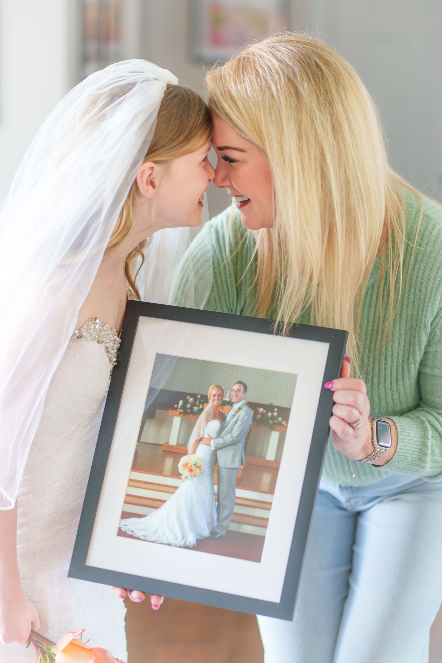 Bride and blonde woman touch foreheads while holding framed wedding photo together in heartfelt moment.