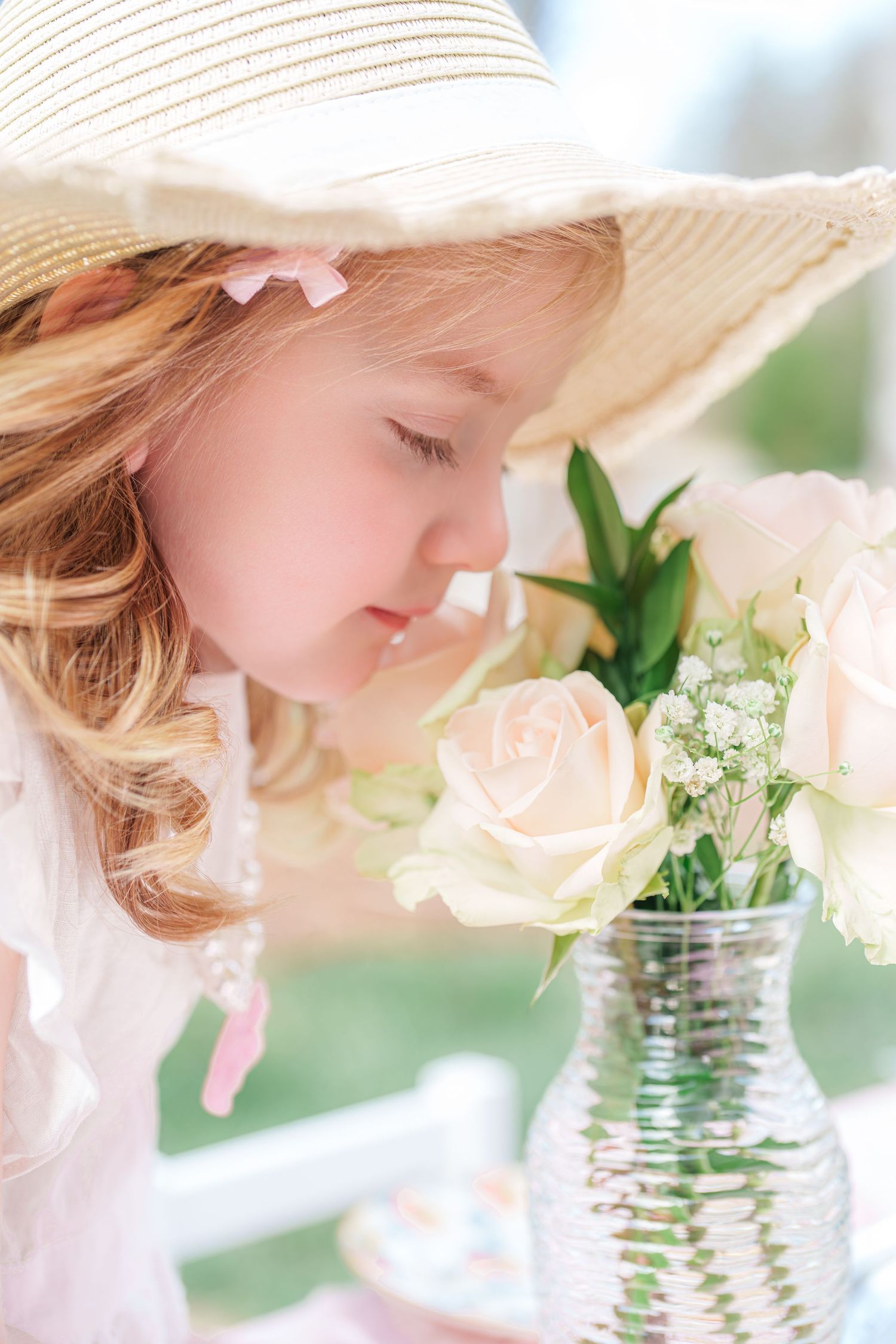A little girl in a wide-brim straw hat smells fresh white roses in a glass vase outdoors.