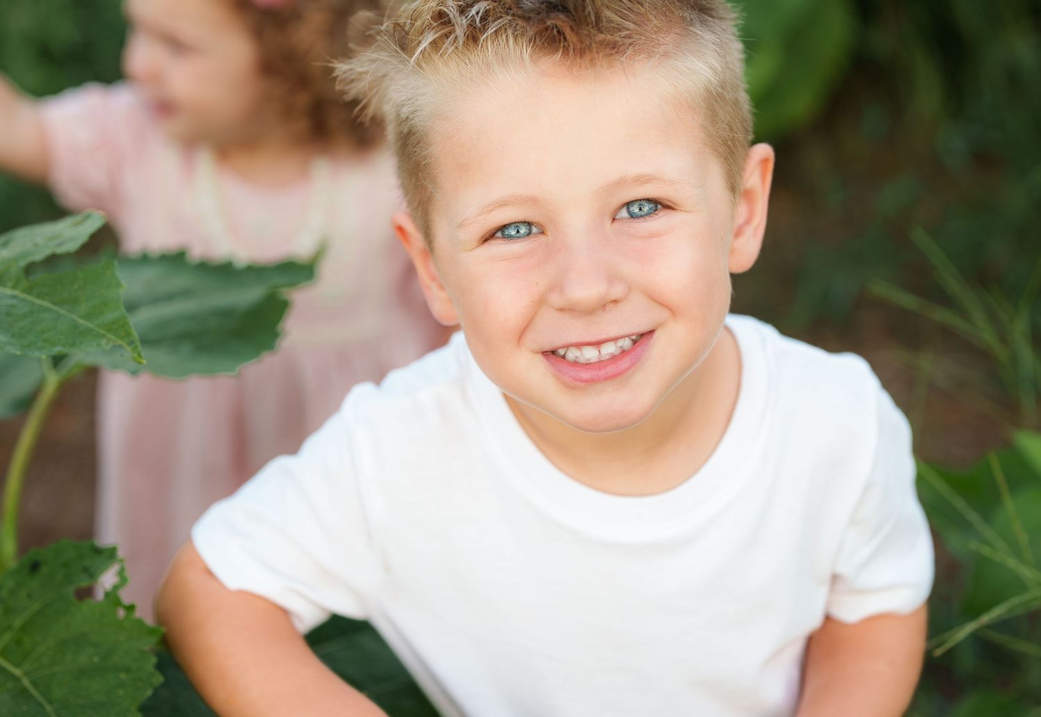Smiling blonde boy with blue eyes in a white t-shirt among green plants outdoors.