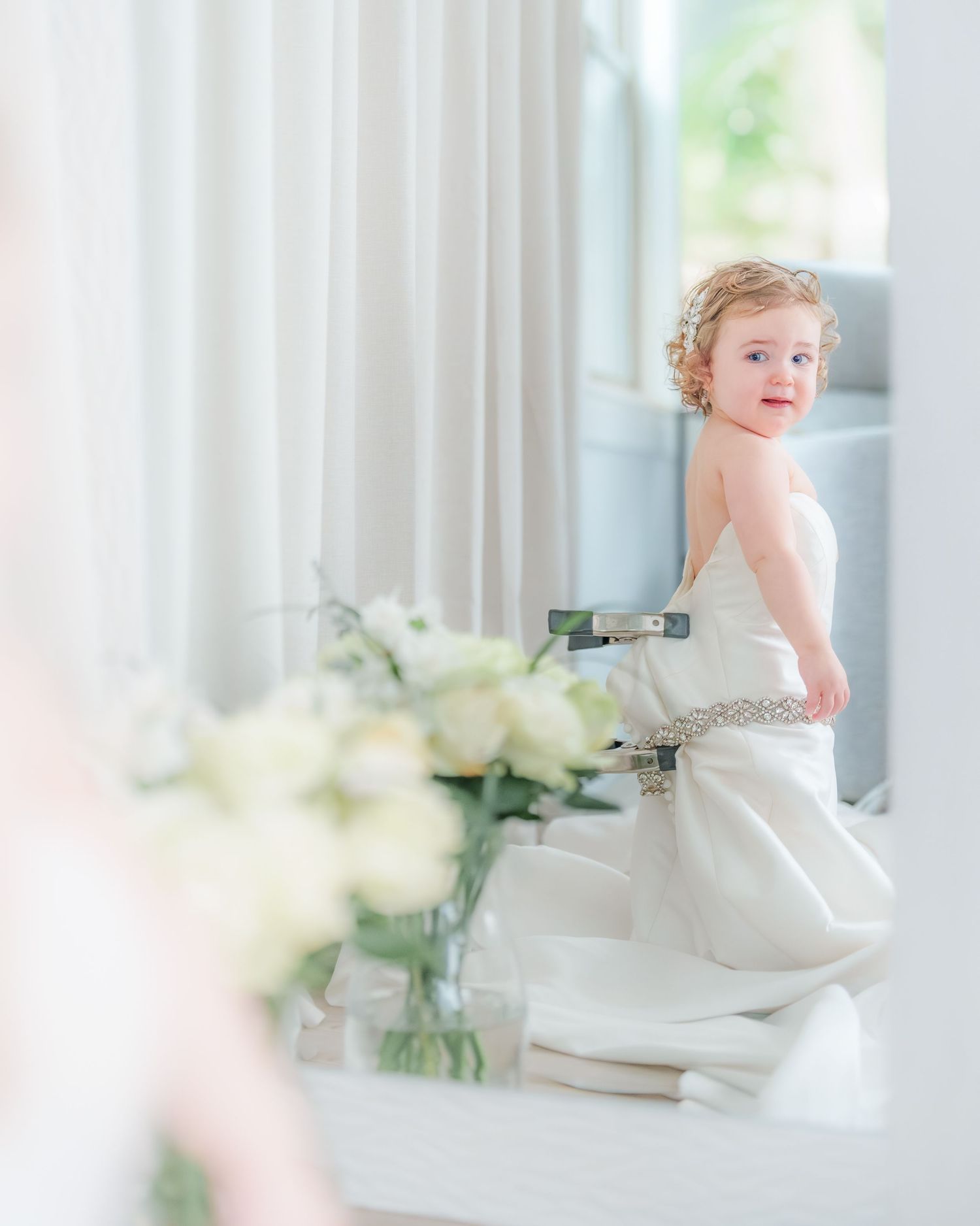 Smiling curly-haired toddler girl in white wedding dress with rhinestone belt near mirror.