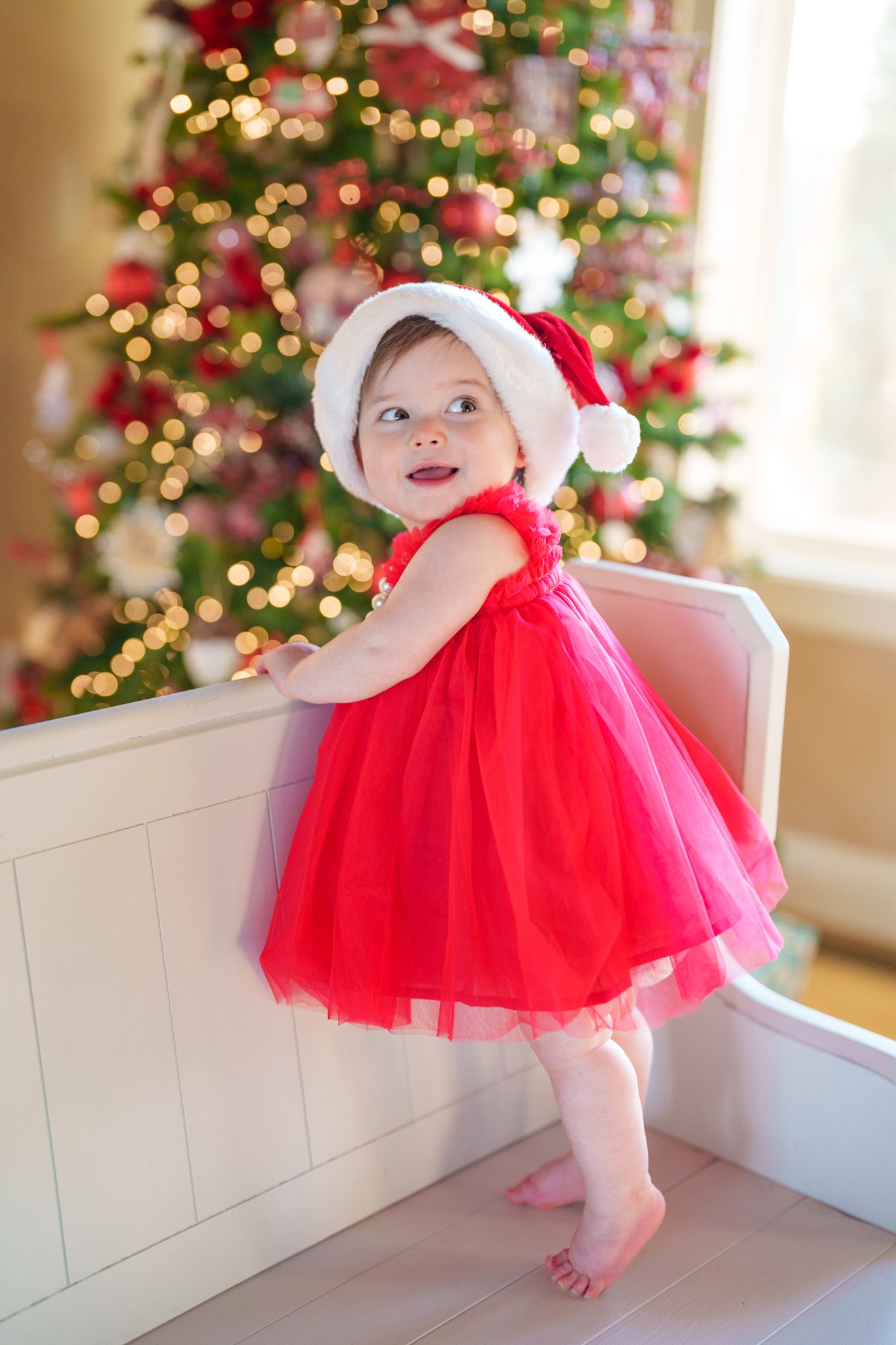 Smiling baby girl in red tulle dress and Santa hat standing by a beautifully decorated Christmas tree.