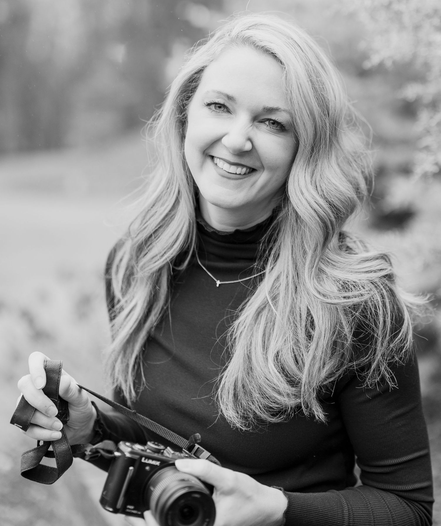 Smiling blonde woman holding a camera outdoors in a black and white portrait photo.