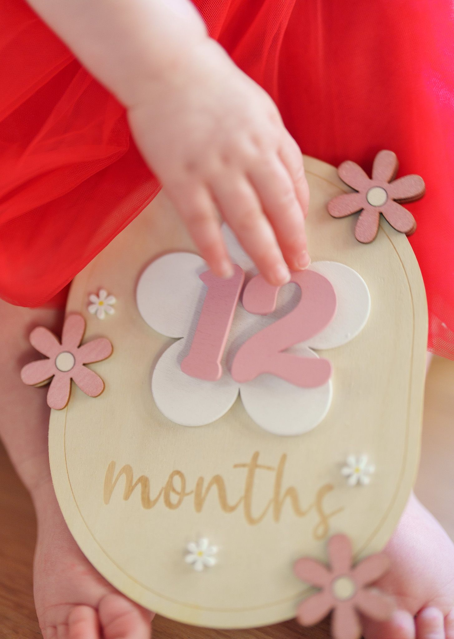 Close-up of baby hands touching a wooden 12 months milestone disc with pink floral decorations.