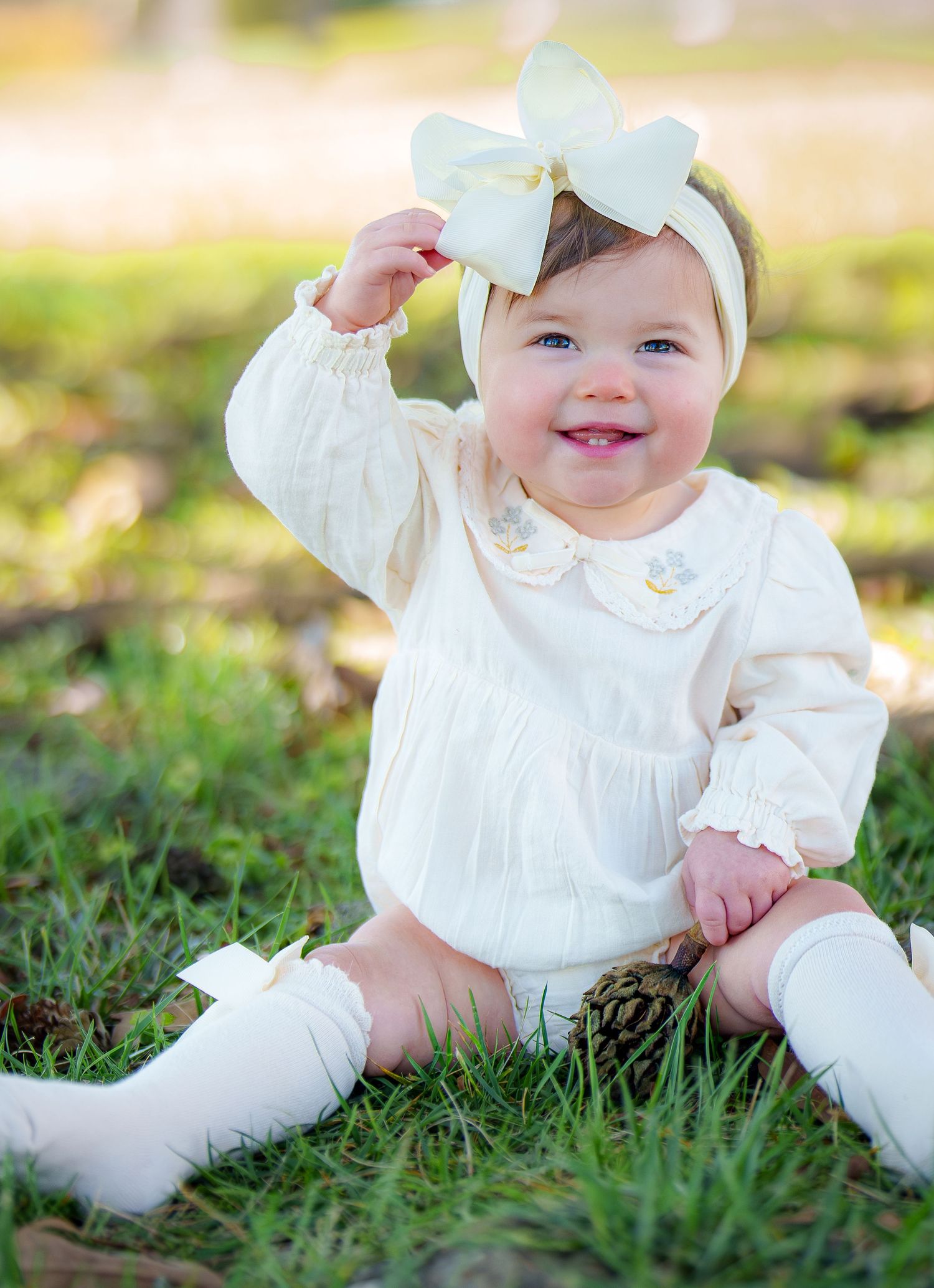 A happy baby girl wearing a cream romper and large bow headband sits on grass, smiling in natural light.