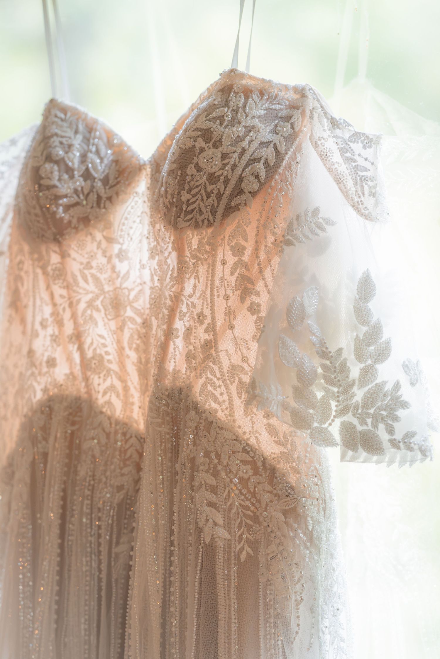 Elegant lace wedding dress with floral embroidery and beading hanging near a window in soft natural light.