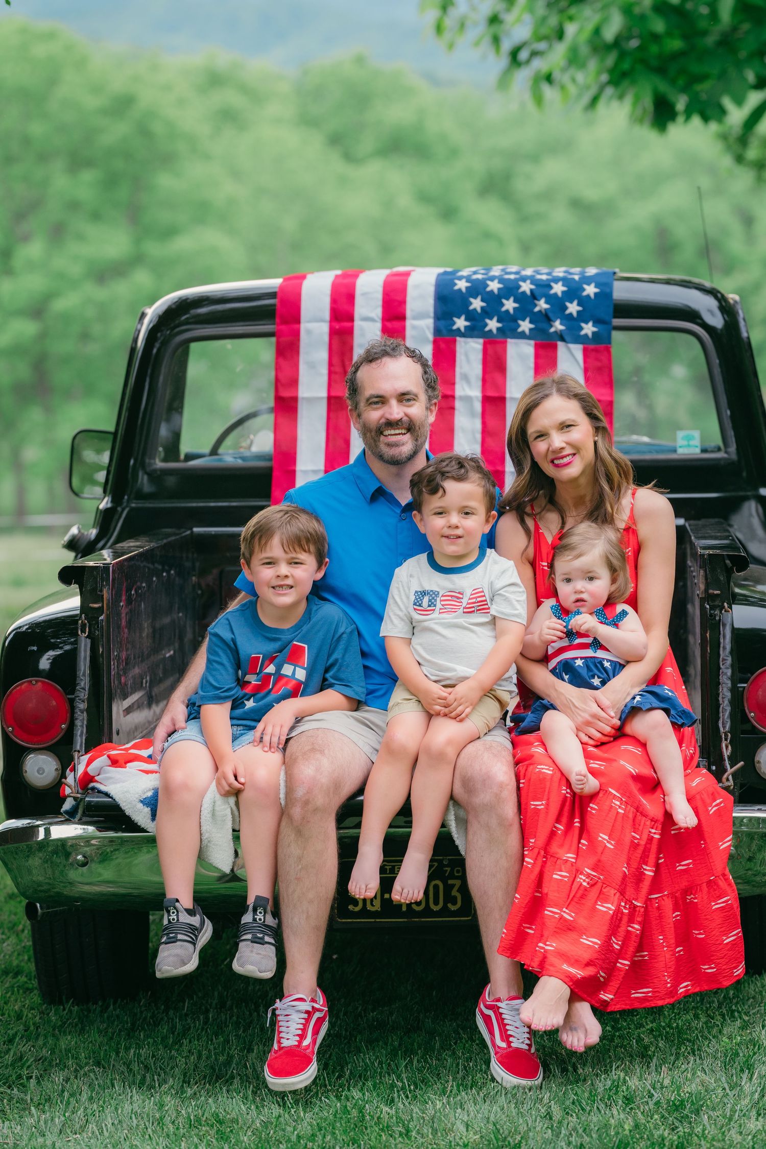 Family of five posing on vintage black truck tailgate with American flag backdrop, dressed in patriotic red, white, and blue outfits.
