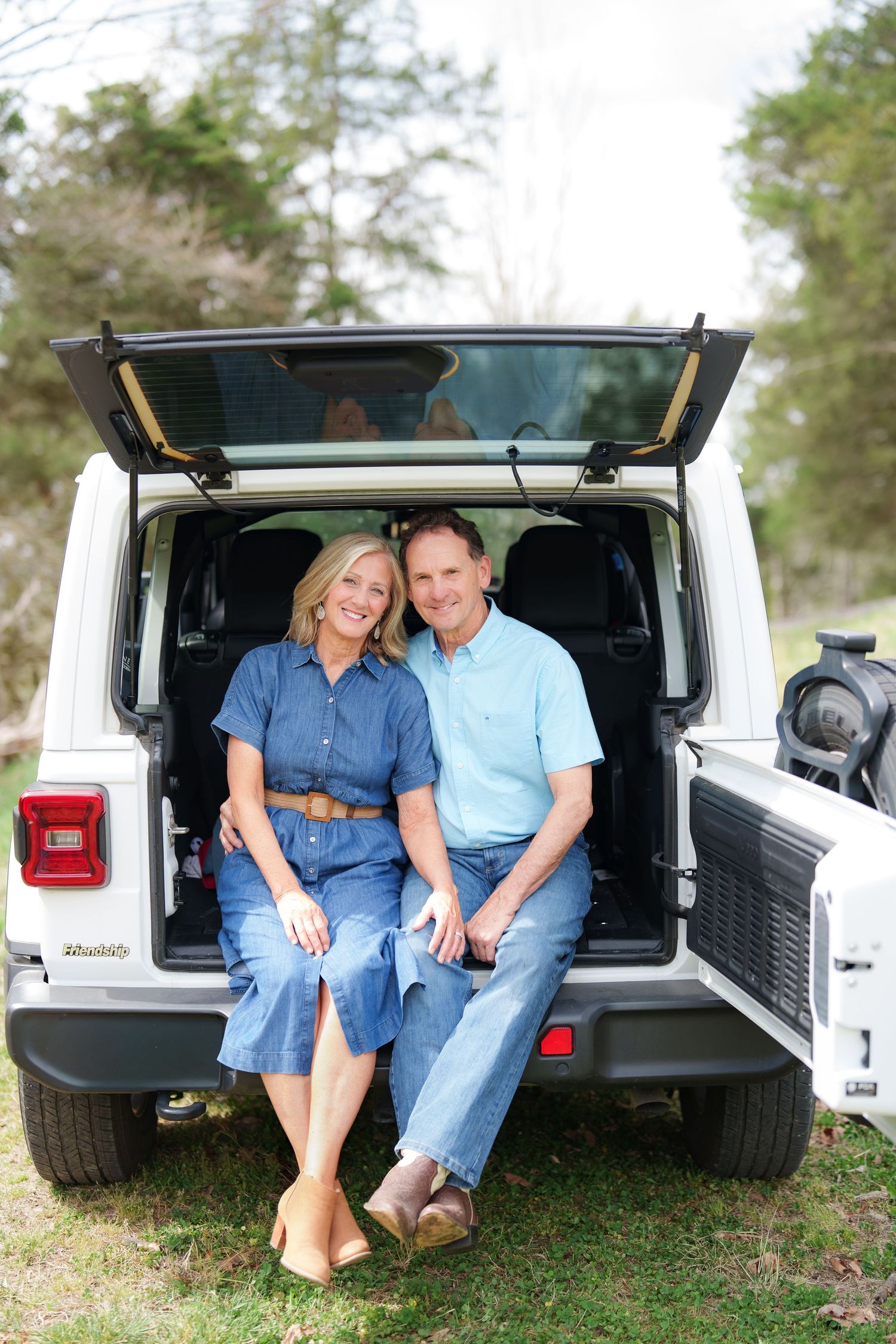 Smiling couple sitting together in the open trunk of a white Jeep parked on a grassy outdoor area.