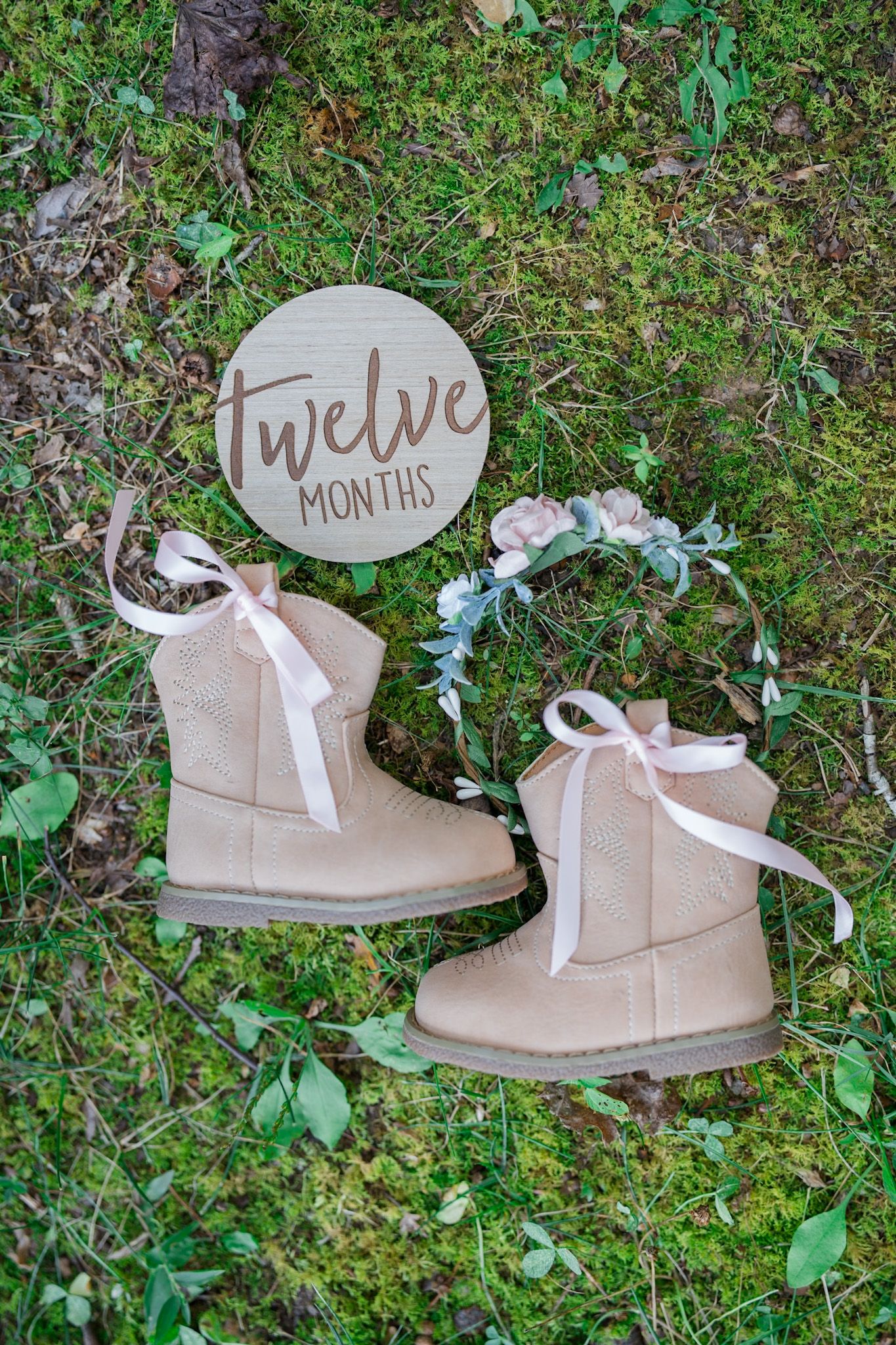 Pink baby cowboy boots with ribbon laces beside a floral crown and twelve months wooden sign on grass.