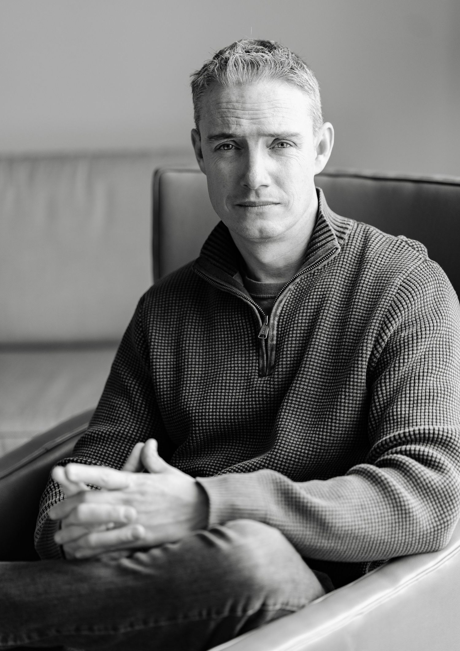 Black and white portrait of a man in a quarter-zip sweater sitting in a modern chair.
