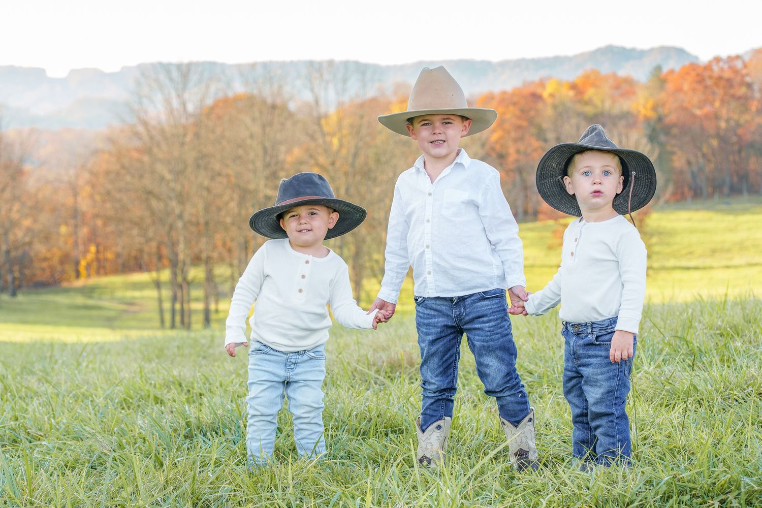 Three young boys in white shirts, jeans, and cowboy hats hold hands in a field with colorful autumn trees.