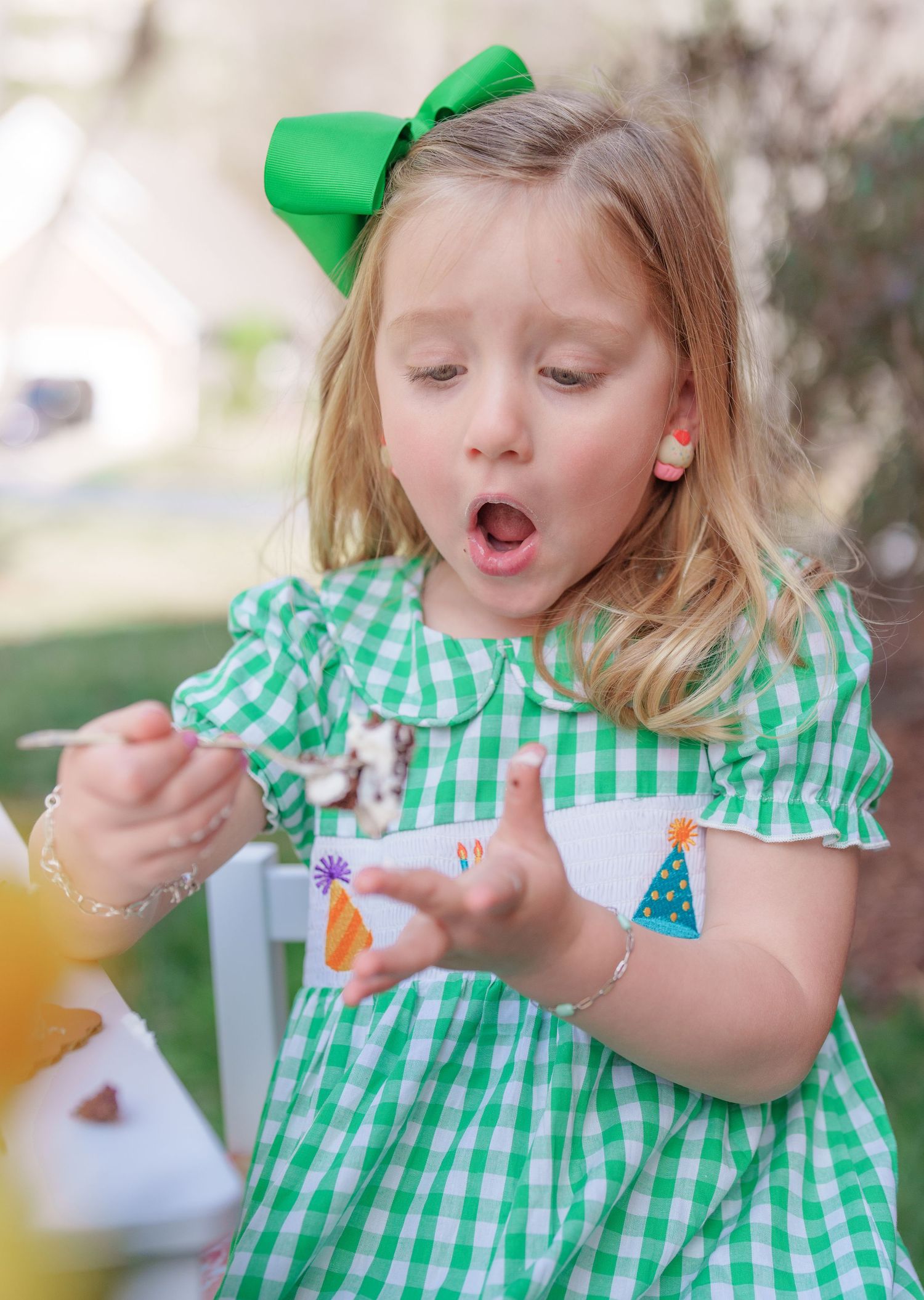 A young girl in a green gingham smocked dress looks surprised while holding a small item at an outdoor tea party.