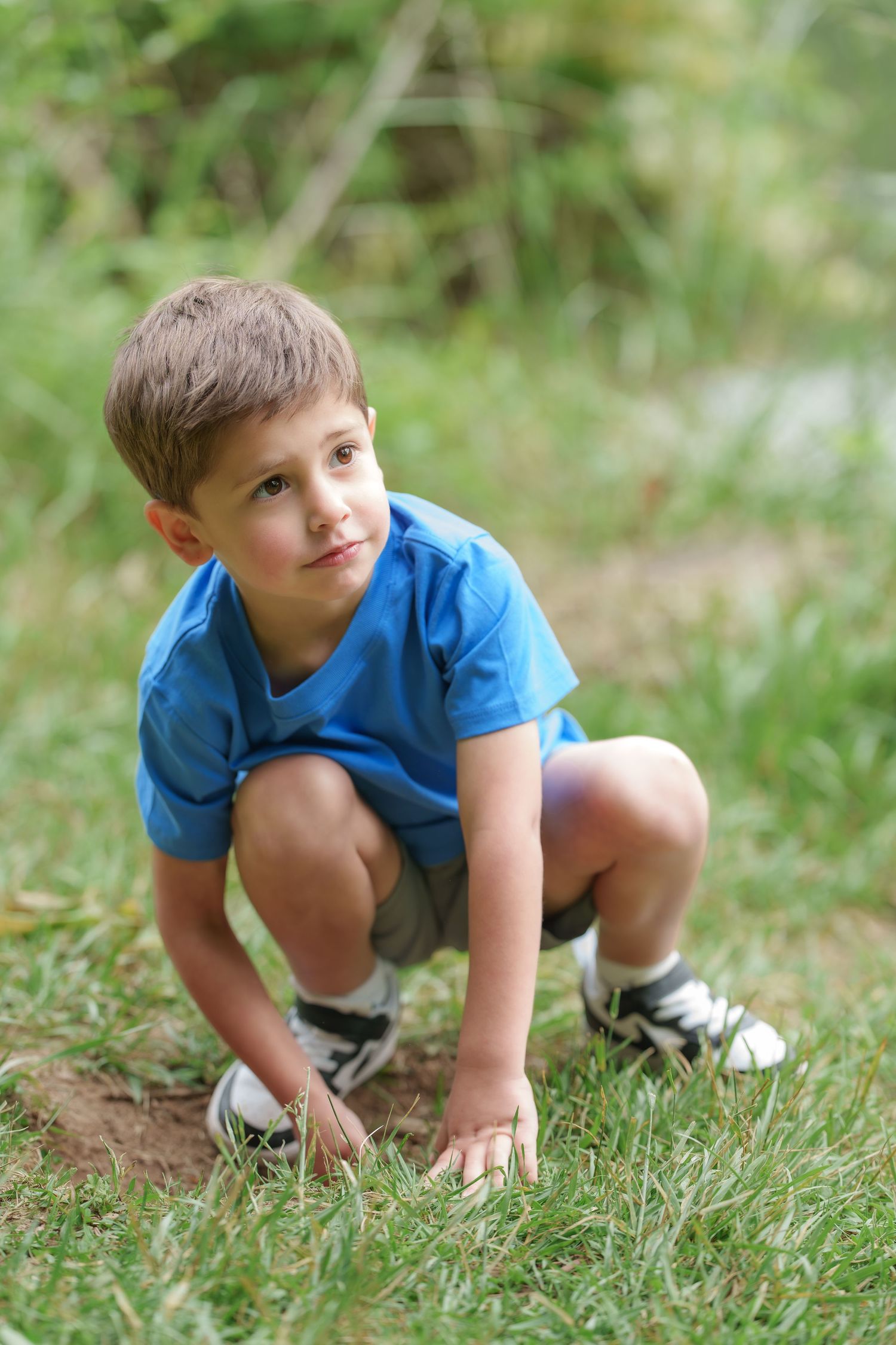 Young boy in blue shirt crouching on grass outdoors, looking up curiously in a natural green setting.