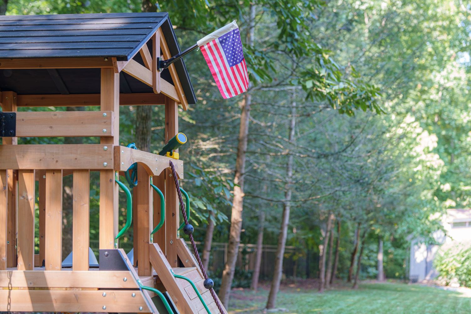 Wooden backyard playset with American flag and green slide surrounded by lush trees on a sunny day.