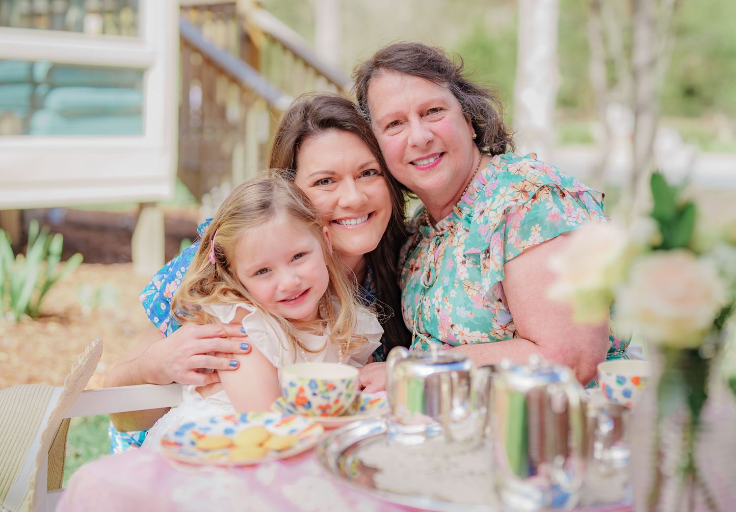 Three generations of women smiling together at a colorful tea party table in a garden setting.
