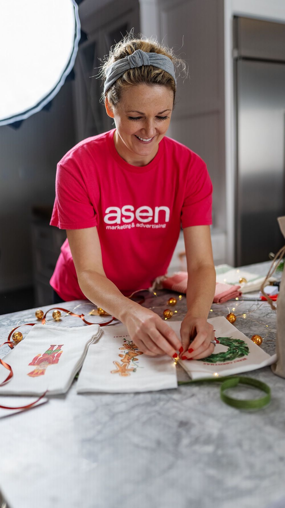 Woman in red Asen marketing t-shirt smiling while decorating Christmas items on a marble table.