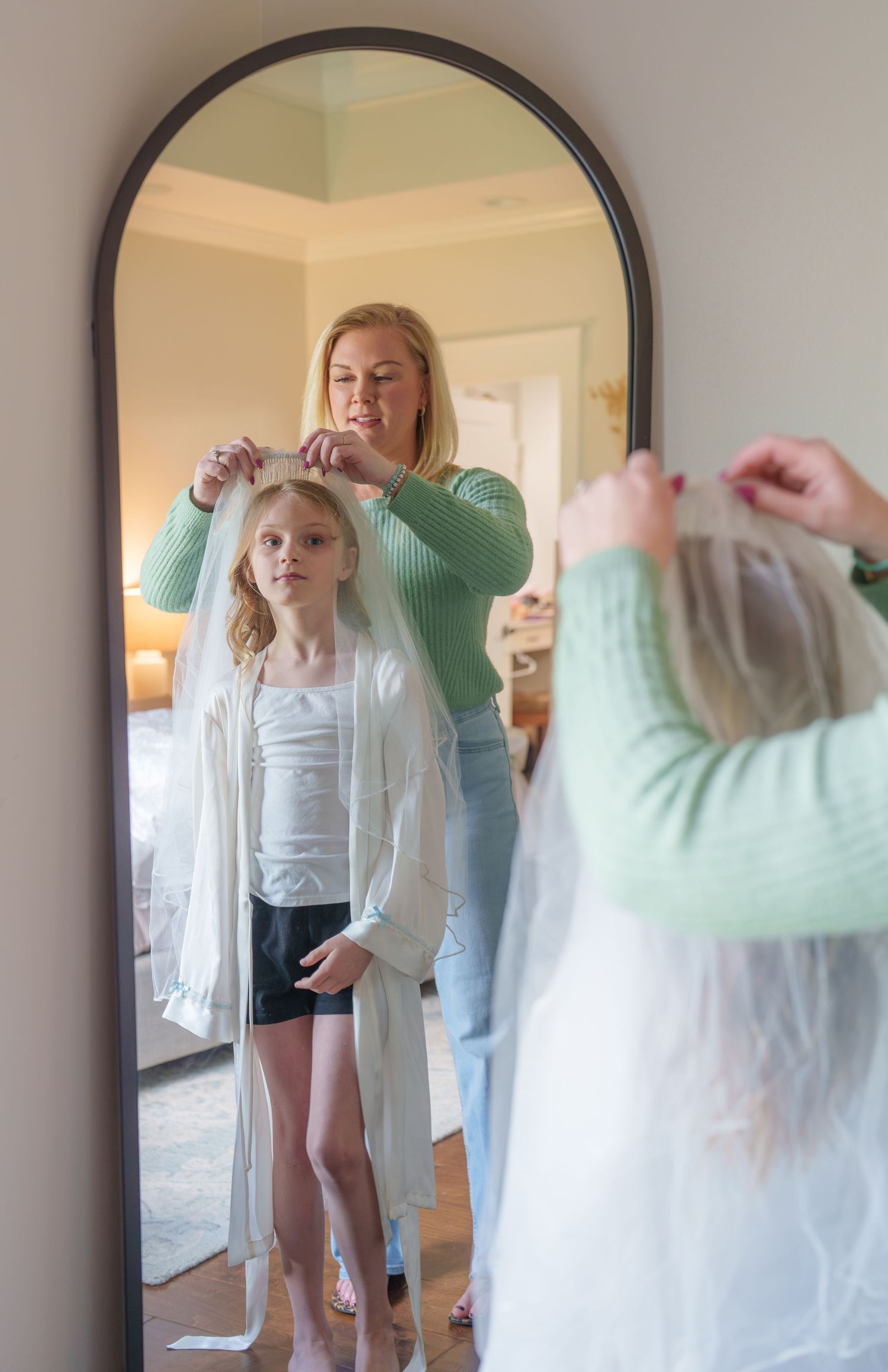 A blonde woman adjusts a wedding veil on a young girl standing before an arched mirror in a bedroom.