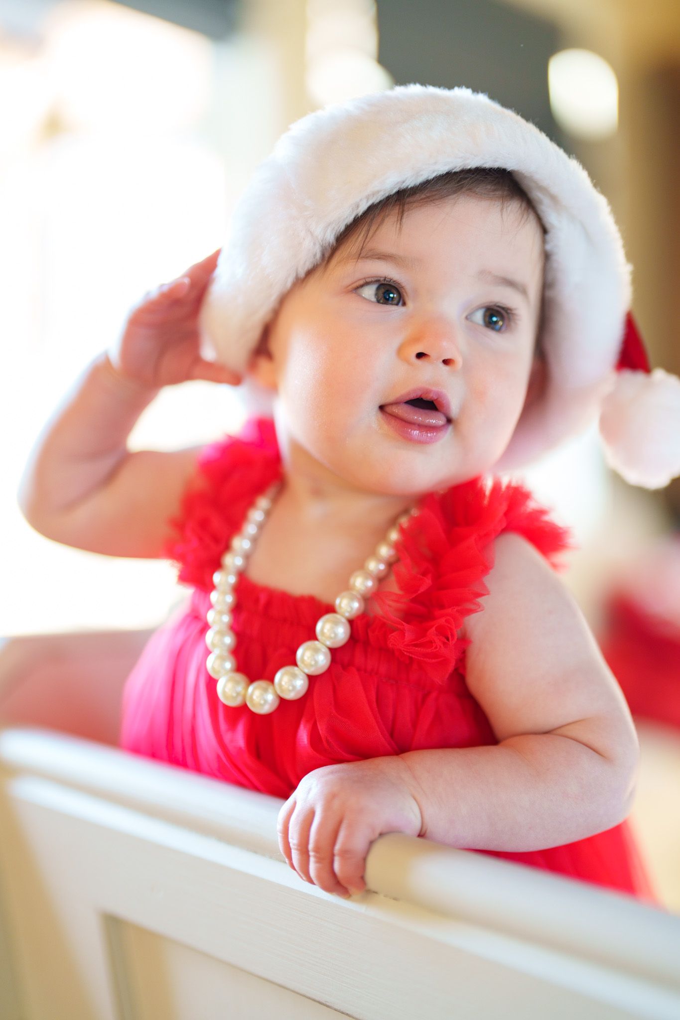 Cute baby girl in red dress and Santa hat wearing pearl necklace stands in white crib.
