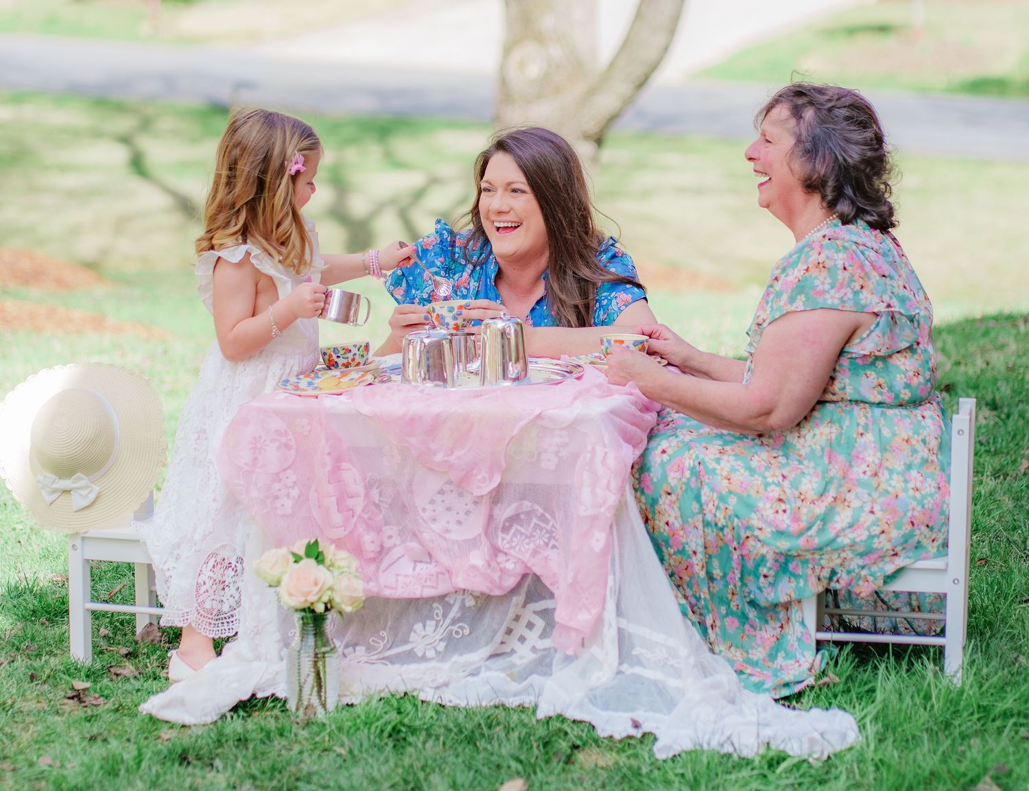 Three generations of women enjoy an outdoor tea party on a pink lace-covered table in a sunny garden.