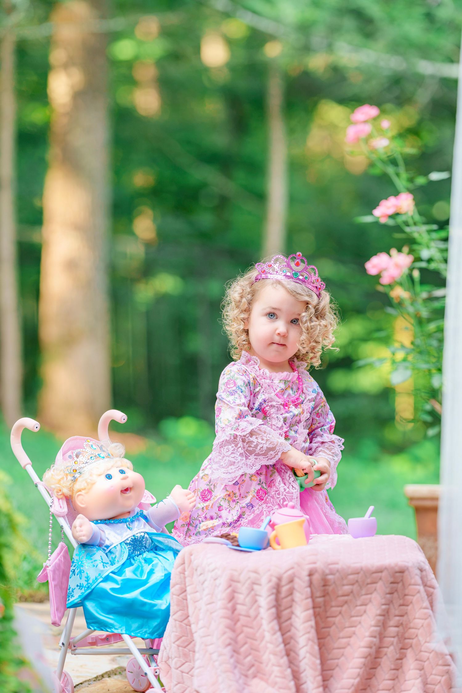 A curly-haired toddler girl in a pink princess dress hosts a outdoor tea party with her doll in a stroller.