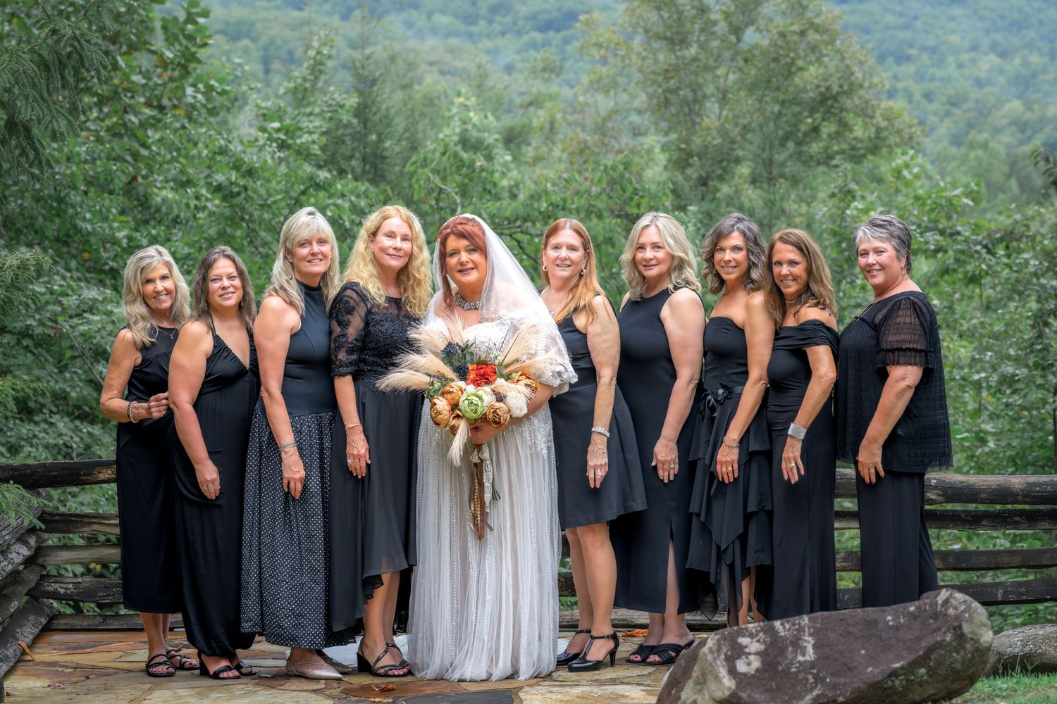 Bride in white gown poses with bridesmaids in black dresses on outdoor deck surrounded by lush green mountains.